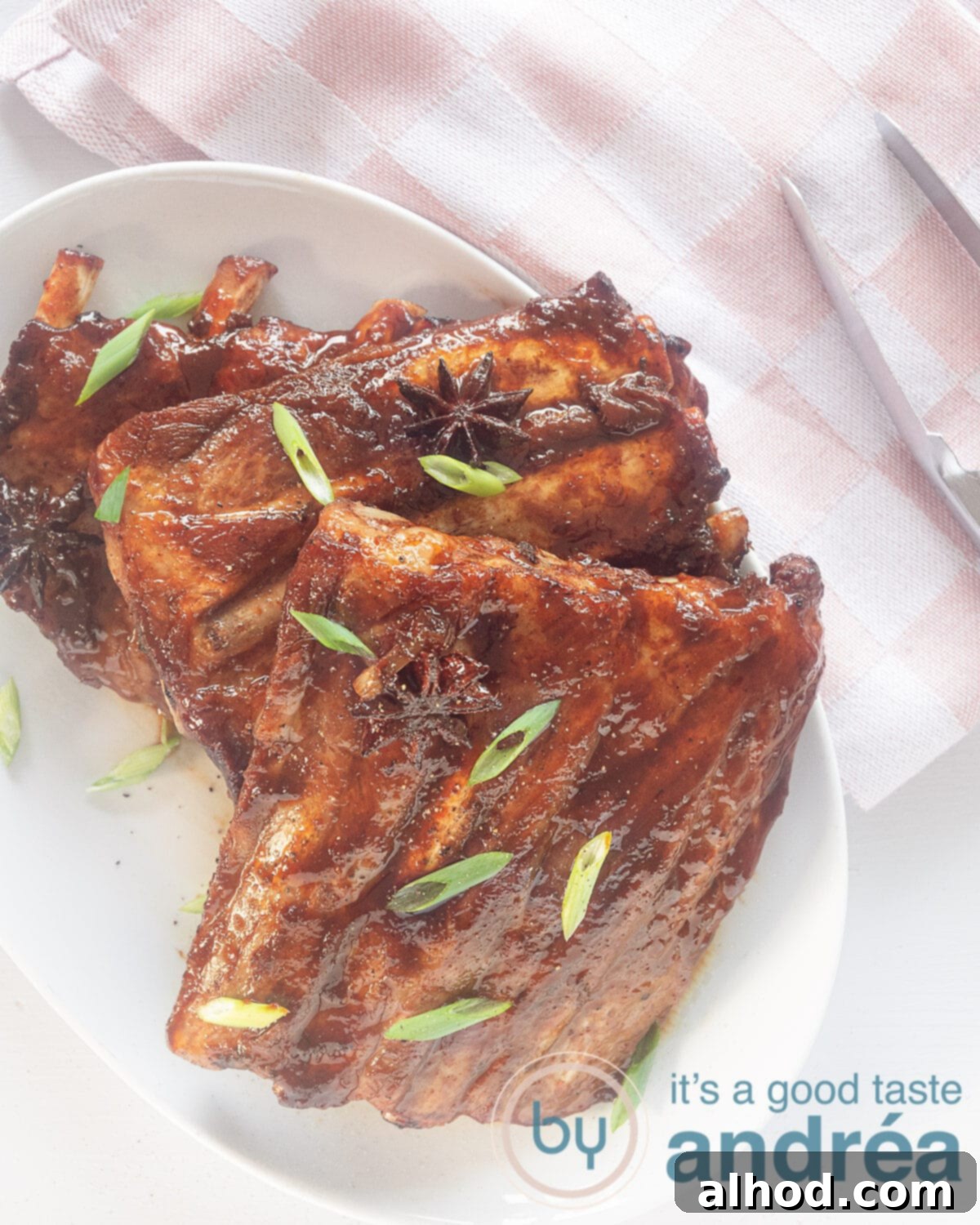 A photo from above, bottom left, a white dish with three sticky ribs sprinkled with spring onion. Top right a pink white checkered tea towel and the end of a clothespin