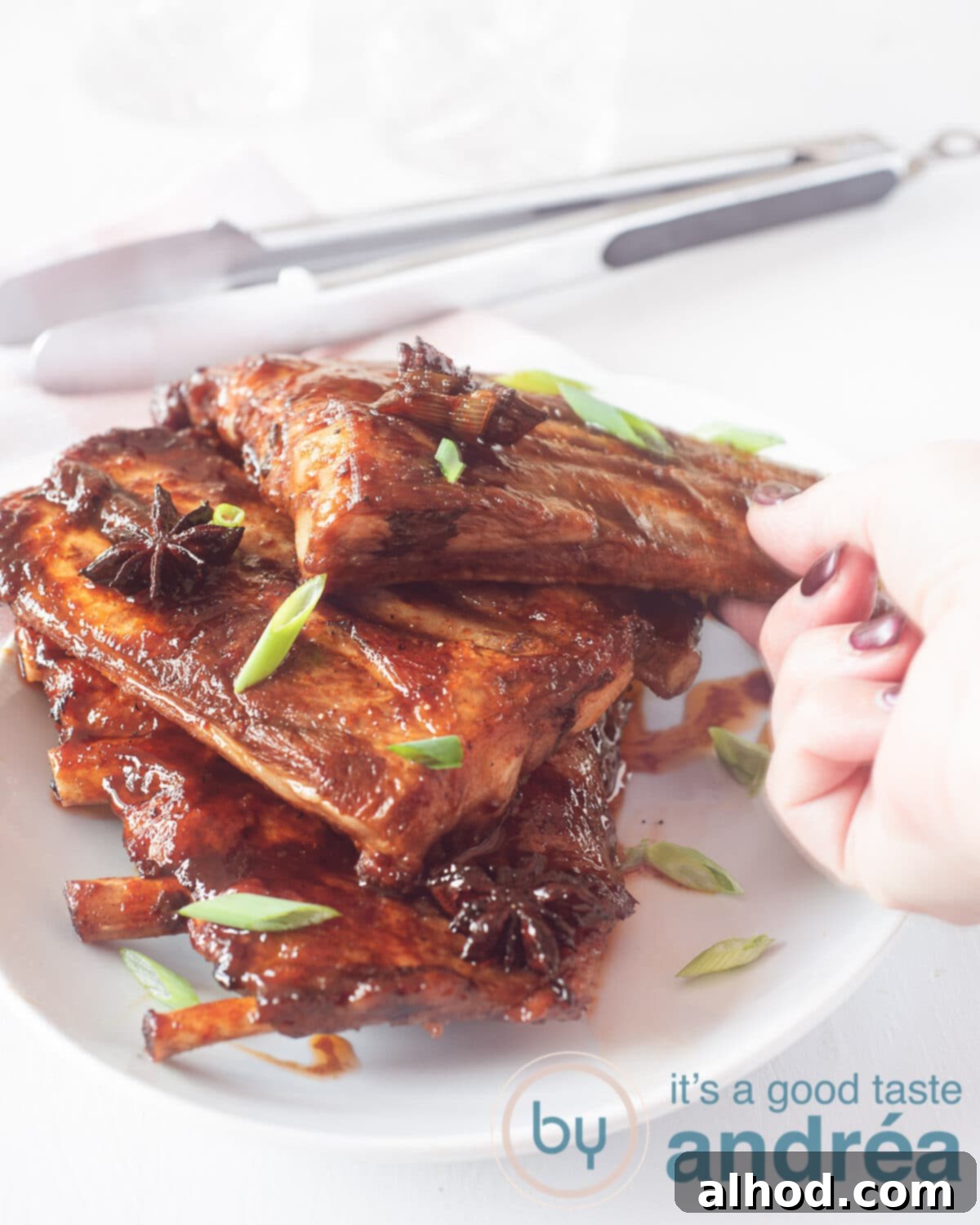 Caramelized Asian Ribs 5 A hand reaches for a perfectly glazed sticky Asian rib from a white dish, garnished with fresh spring onions, against a clean white background. A rustic wooden clothespin rests in the soft-focus background, adding a homely touch.