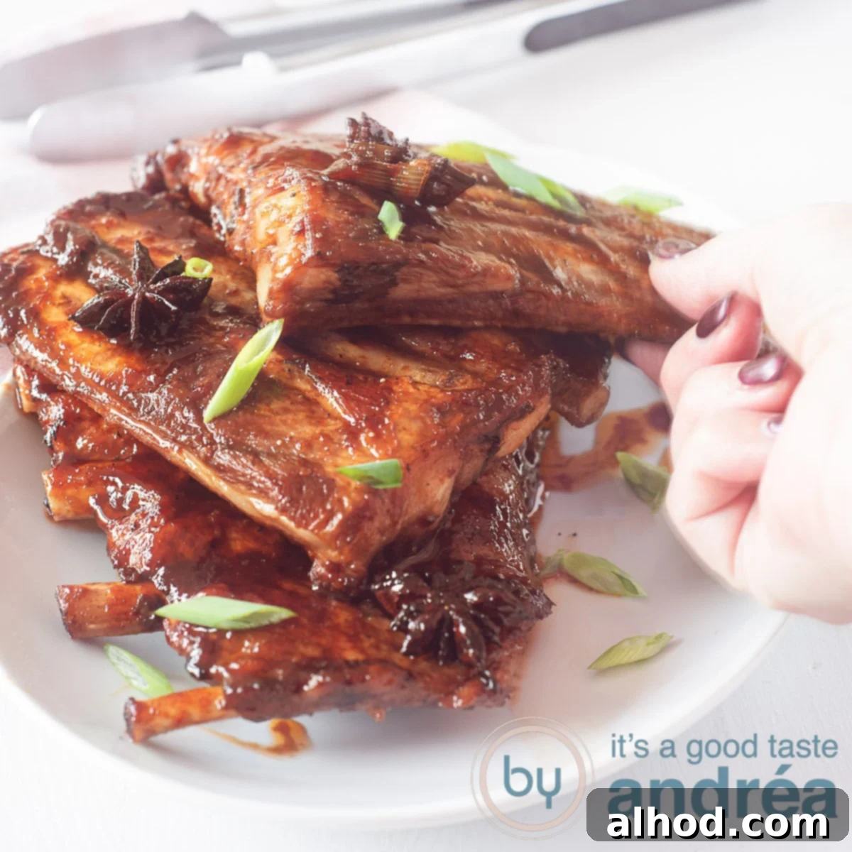 Caramelized Asian Ribs 6 A square photo featuring a tempting stack of sticky spareribs, garnished generously with fresh spring onions. A hand is gently reaching for one of the succulent ribs from the inviting bowl.