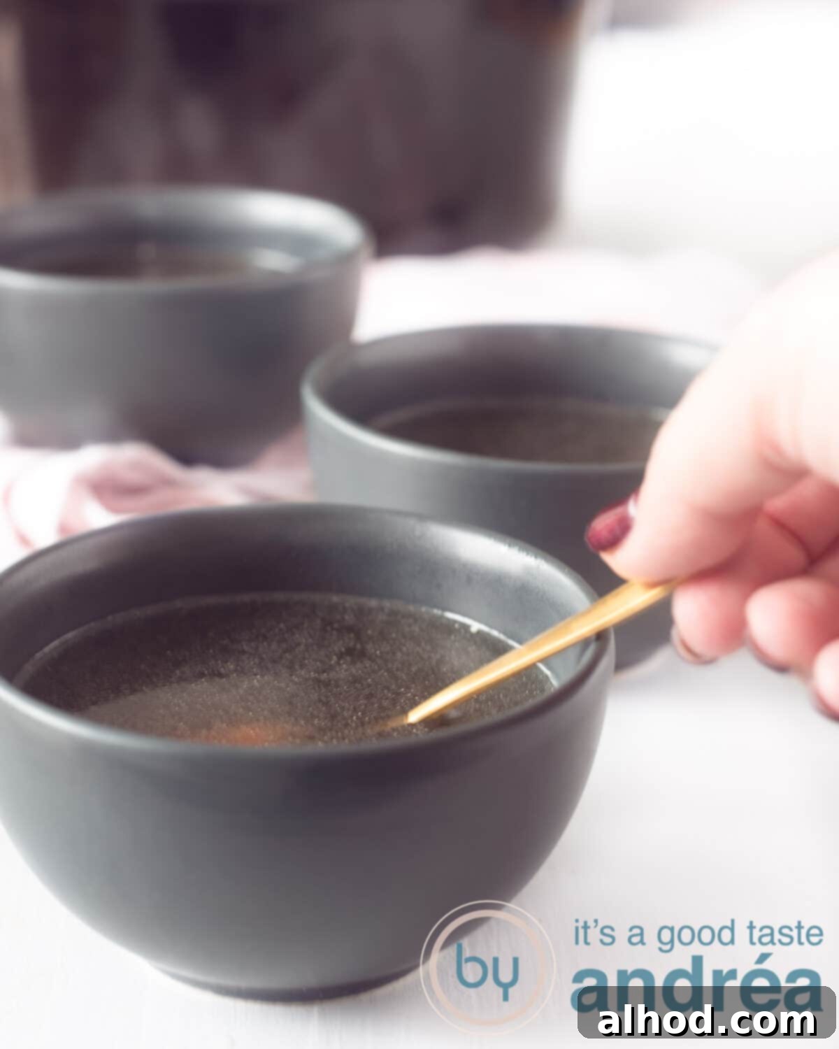 Nourishing Slow Cooker Chicken Bone Broth 5 Three gray dishes on a white background filled with homemade chicken stock. A golden spoon is in one, with a hand holding the spoon, ready to taste. A slow cooker is visible in the background, hinting at the ease of preparation.