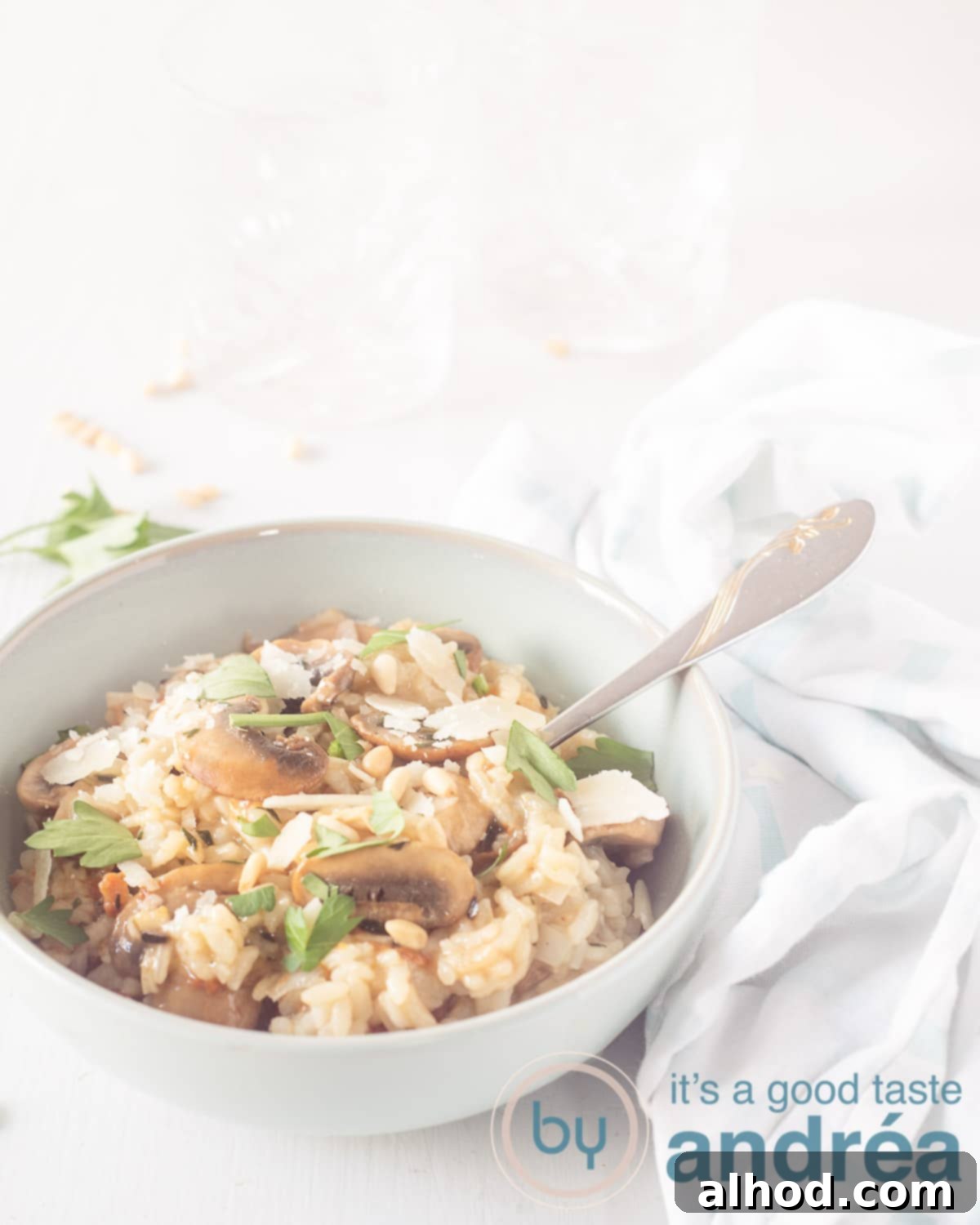 Earthy Mushroom Risotto 2 A blue-green dish at the bottom left with a spoon, filled with risotto, chestnut mushroom, parsley and pine nuts. On a white background. A white and blue tea towel on the right.