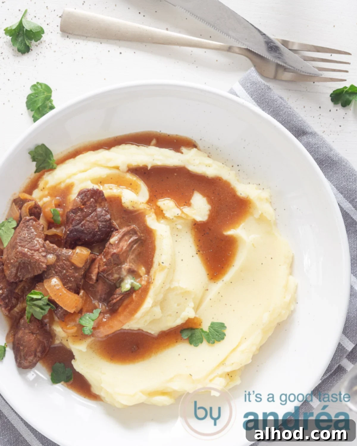 Classic Dutch Hachee Beef and Onion Stew 2 A photo from above of a white plate filled with fluffy mashed potatoes, generous pieces of tender hachee (Dutch beef and onion stew) in a rich, dark gravy. Fresh green parsley garnishes the plate. The meal rests on a clean white background, with a golden fork and knife positioned above, ready to enjoy.