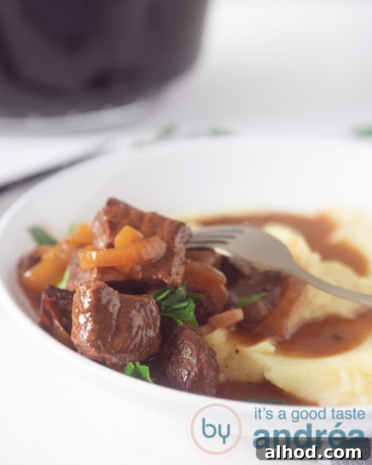 Classic Dutch Hachee Beef and Onion Stew 5 A close-up shot of a half-filled white deep plate, showcasing creamy mashed potatoes on one side and generous portions of hachee (Dutch beef and onion stew) with visible onions and a rich, glossy gravy. A golden fork is poised, piercing a succulent piece of beef. In the softly blurred background, a slow cooker is visible, hinting at the ease of preparation. This image perfectly captures the comfort and deliciousness of a homemade Dutch meal.