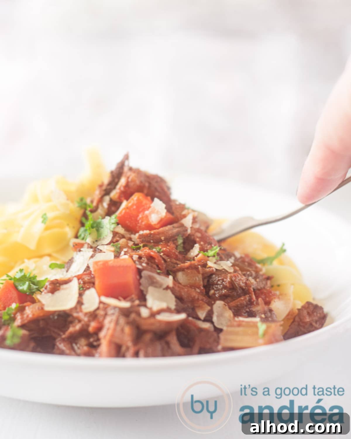 Tender Slow Cooker Beef Ragu with Pasta 5 A white plate filled with Beef Ragu with carrot and Parmesan cheese and papardelle. On a white background. A fork pierces a piece of meat