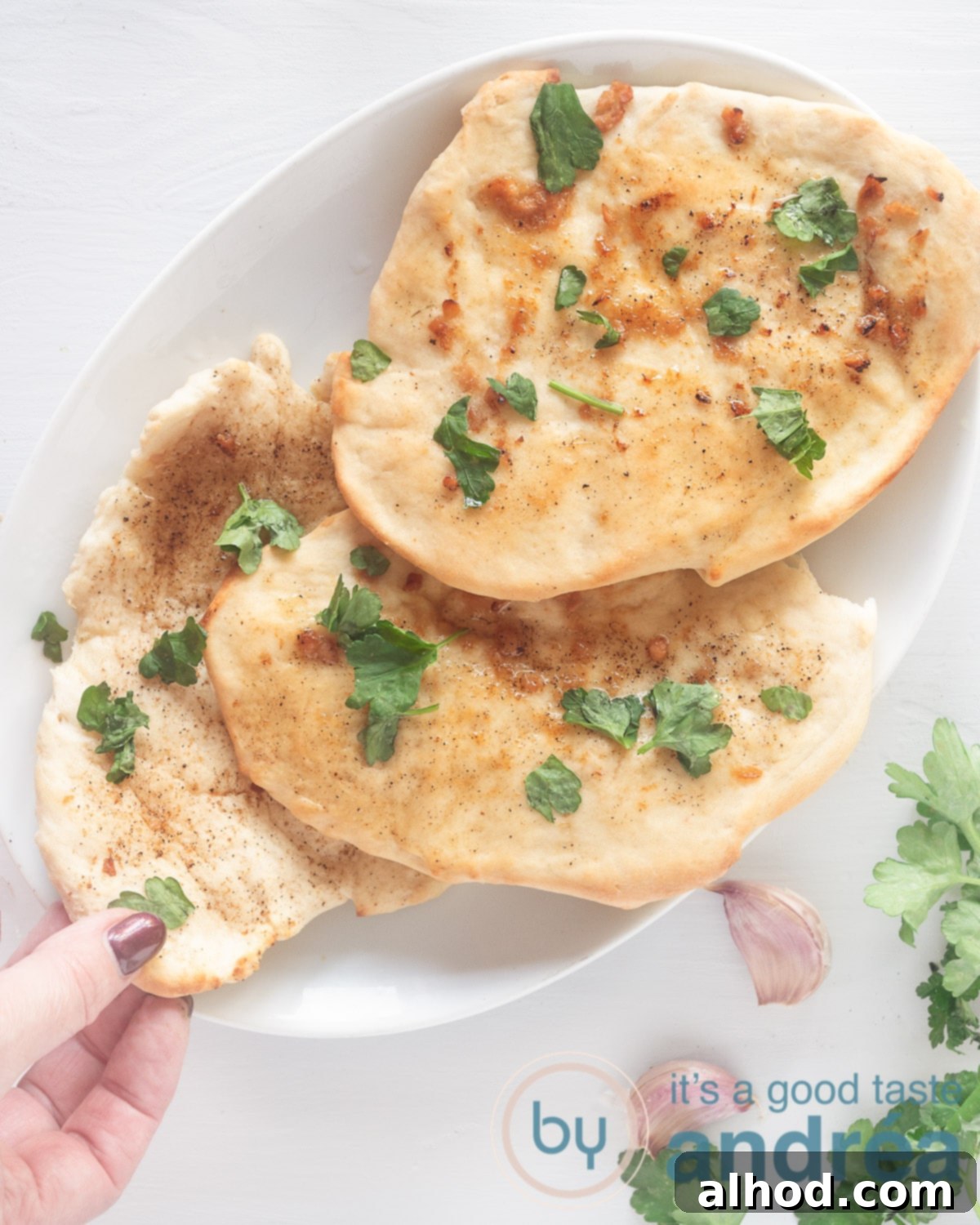Buttery Garlic Herb Naan 2 A photo from above, a white bowl with three naans sprinkled with parsley. A hand takes a loaf of bread. A white background, at the bottom right parsley and garlic