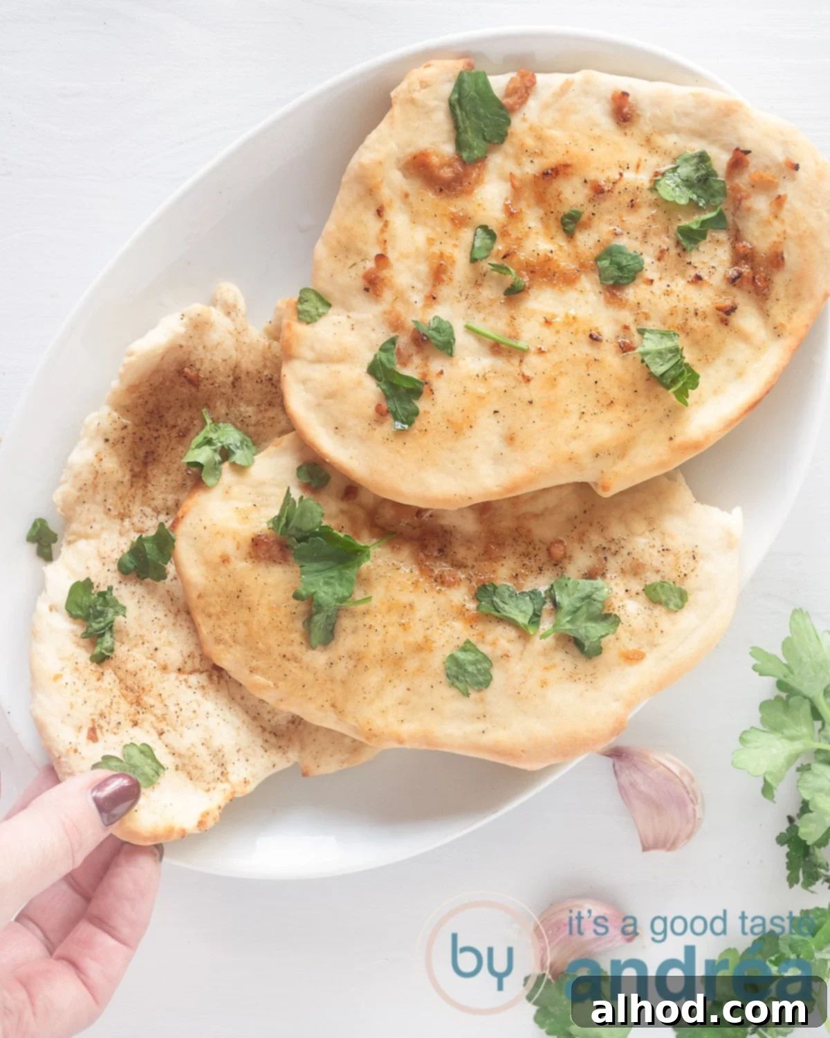 Buttery Garlic Herb Naan 2 A photo from above, a white bowl with three naans sprinkled with parsley. A hand takes a loaf of bread. A white background, at the bottom right parsley and garlic