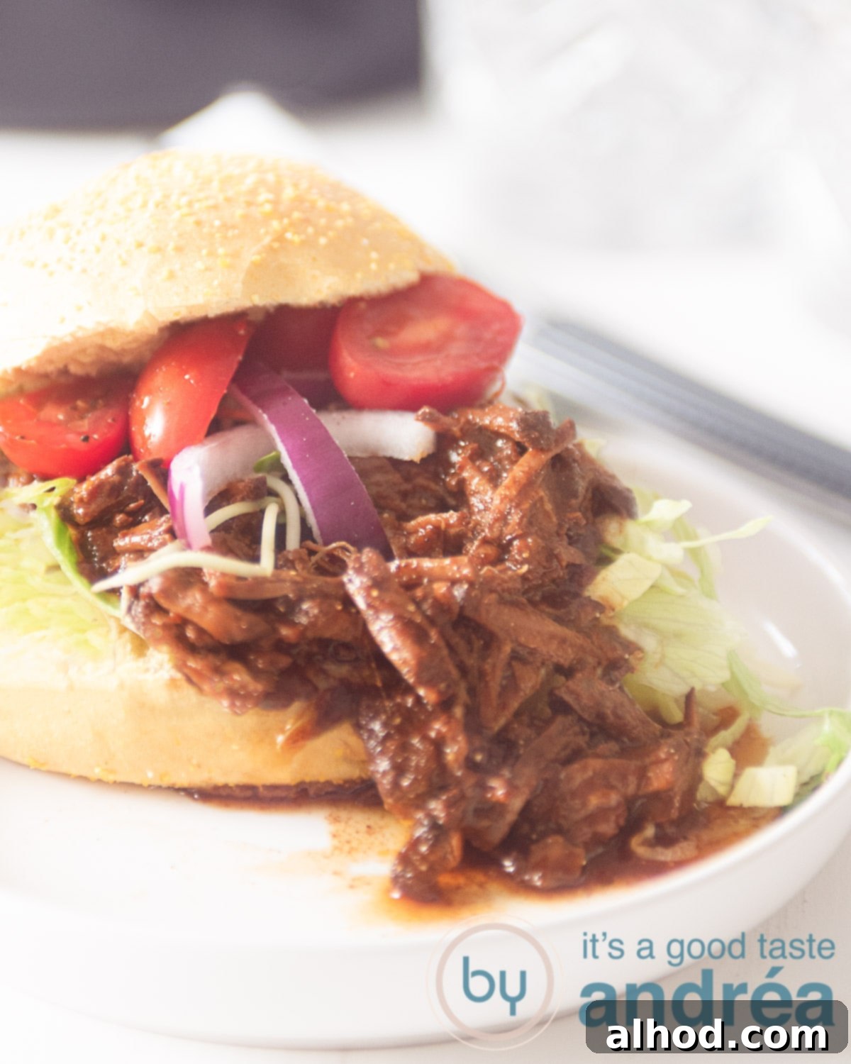 Sticky Asian Slow Cooker Beef 5 A white plate with a sandwich draped with Pulled Asian beef, red onion, tomato and iceberg lettuce. The cap on. On a white background