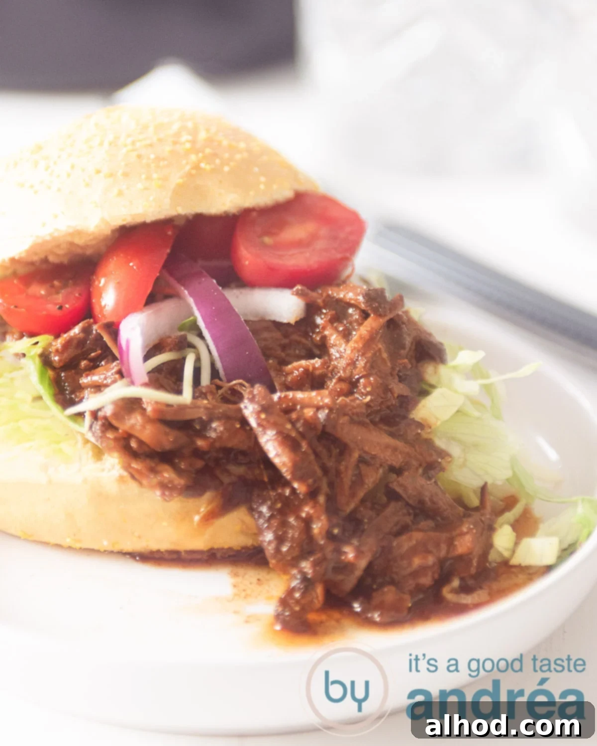 Umami-Rich Asian Slow Cooker Beef 5 A white plate with a sandwich draped with Pulled Asian beef, red onion, tomato and iceberg lettuce. The cap on. On a white background