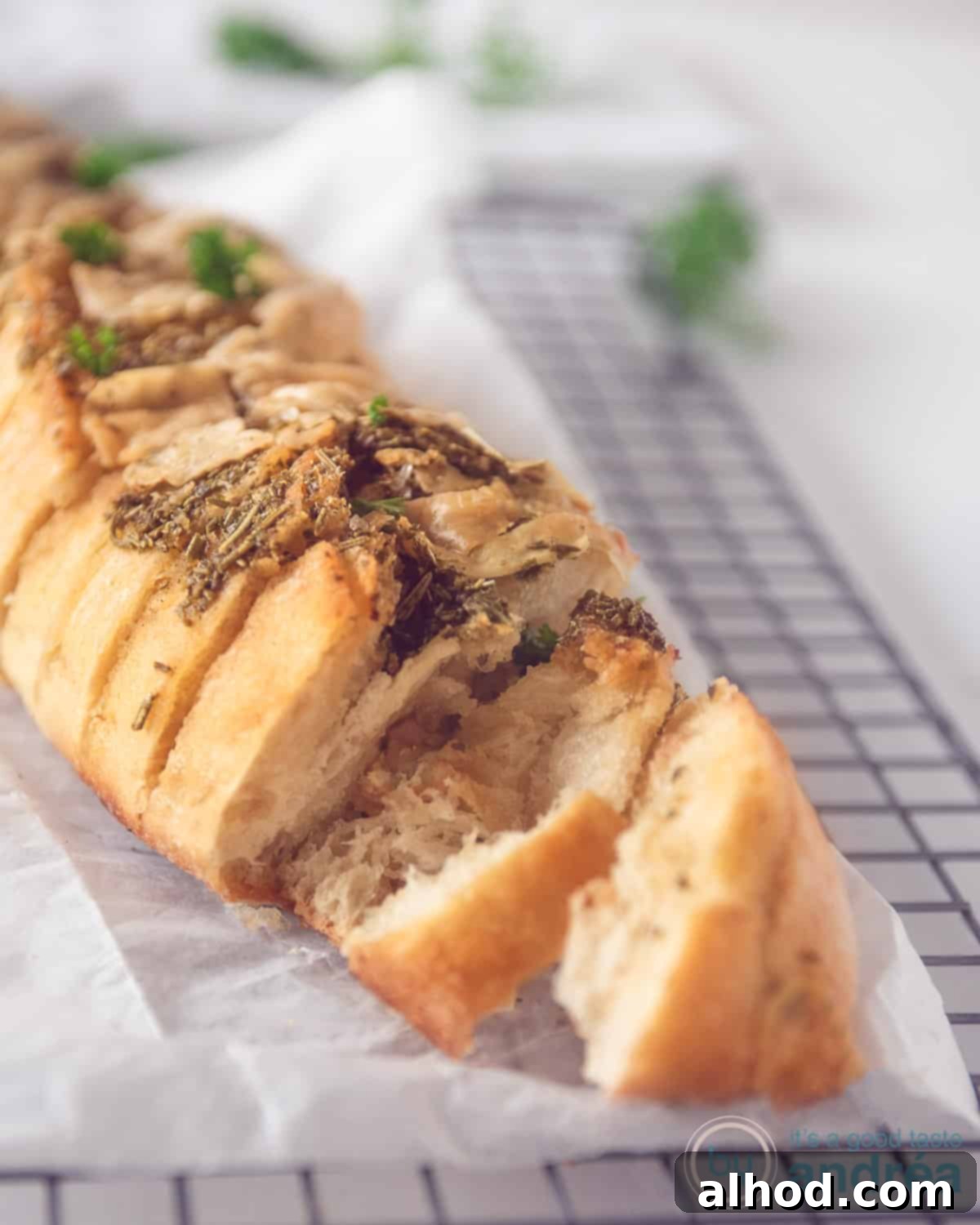 Herbed Garlic Baguette 2 A golden-brown baguette generously topped with fresh herbs and garlic, resting on baking paper. Two slices are neatly cut, revealing the aromatic, cheesy interior.