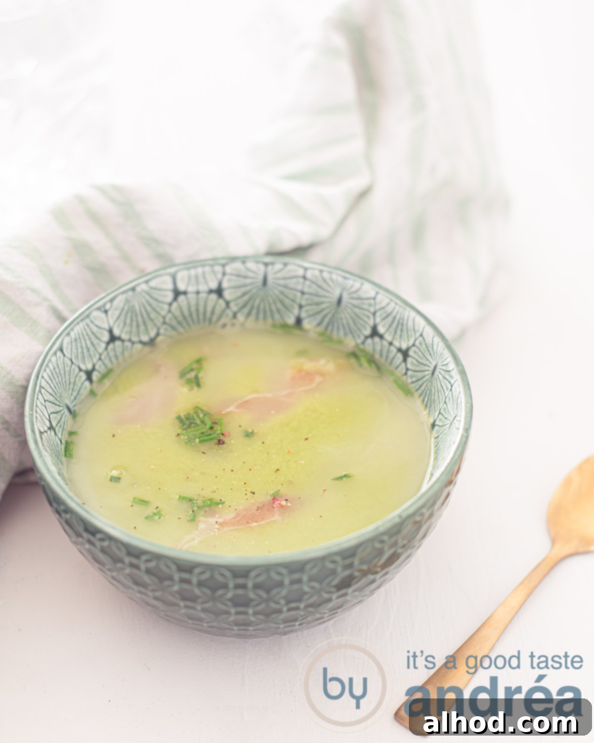 Vibrant Fresh Pea Soup 2 A green bowl filled with vibrant fresh pea soup, garnished with crispy prosciutto strips and fresh chives, set on a clean white background. A golden spoon rests on the right, and a stylish green and white striped towel is on the left.