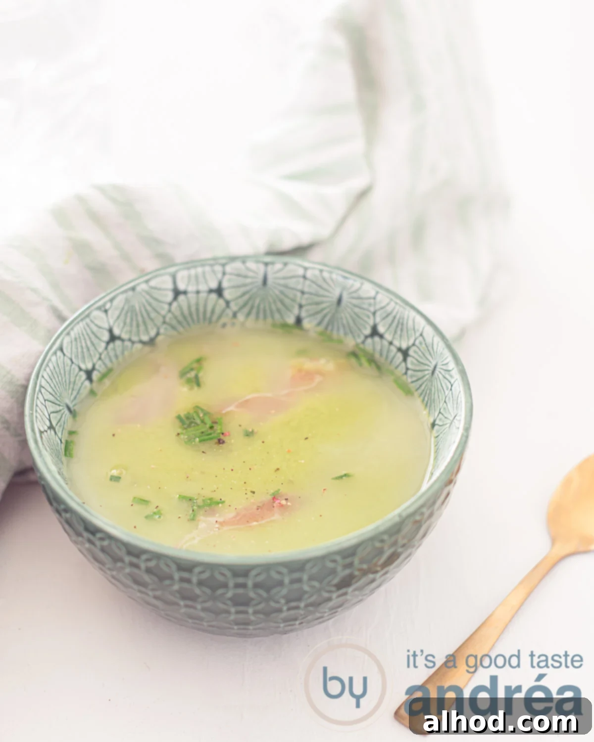 Garden Fresh Pea Soup 2 A green bowl filled with creamy fresh pea soup, garnished with crispy prosciutto and fresh chives, set on a clean white background with a golden spoon and a green and white striped towel.