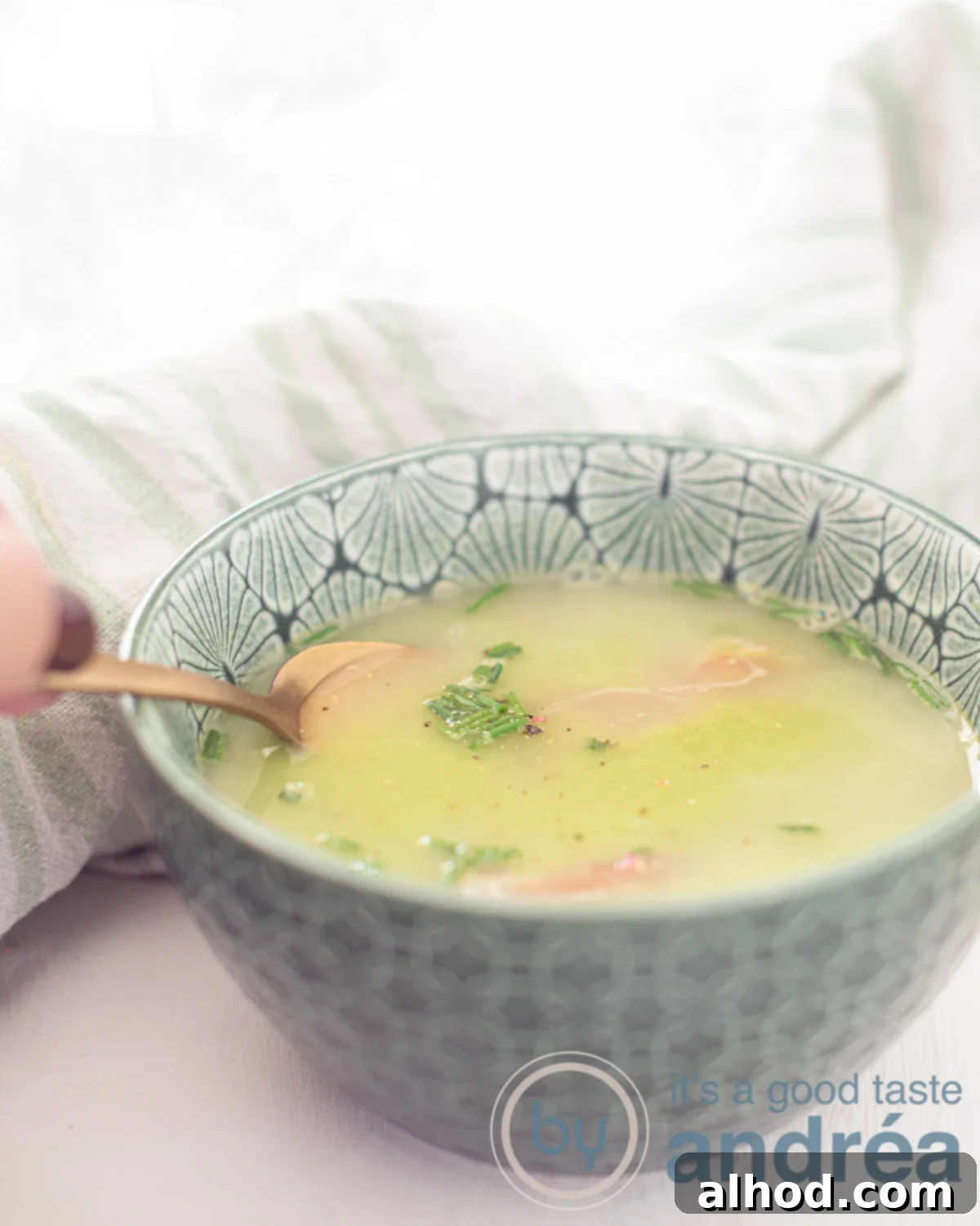 Vibrant Fresh Pea Soup 5 A close-up of a green bowl filled with creamy pea soup, garnished with fresh chives and delicate strips of ham. A hand is dipping a spoon into the soup, with a soft white background and a green and white tea towel in the background, suggesting a cozy meal.