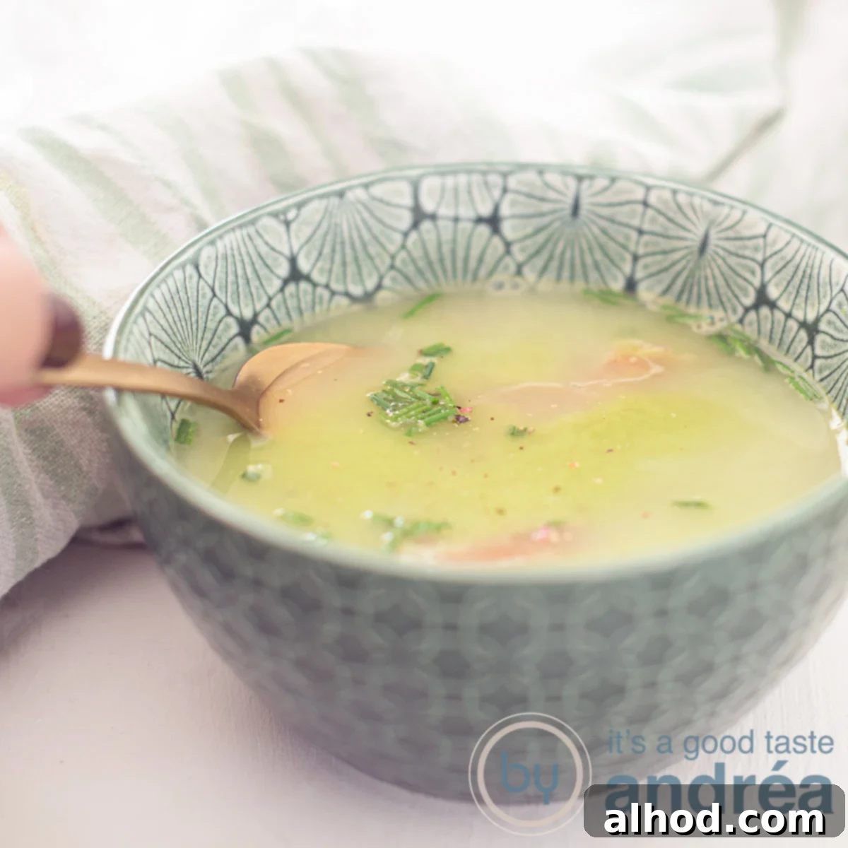 Vibrant Fresh Pea Soup 6 A square photo with a green bowl filled with fresh pea soup, garnished with Serrano ham, cream, and chives. A hand reaches for a bite, all on a white background.