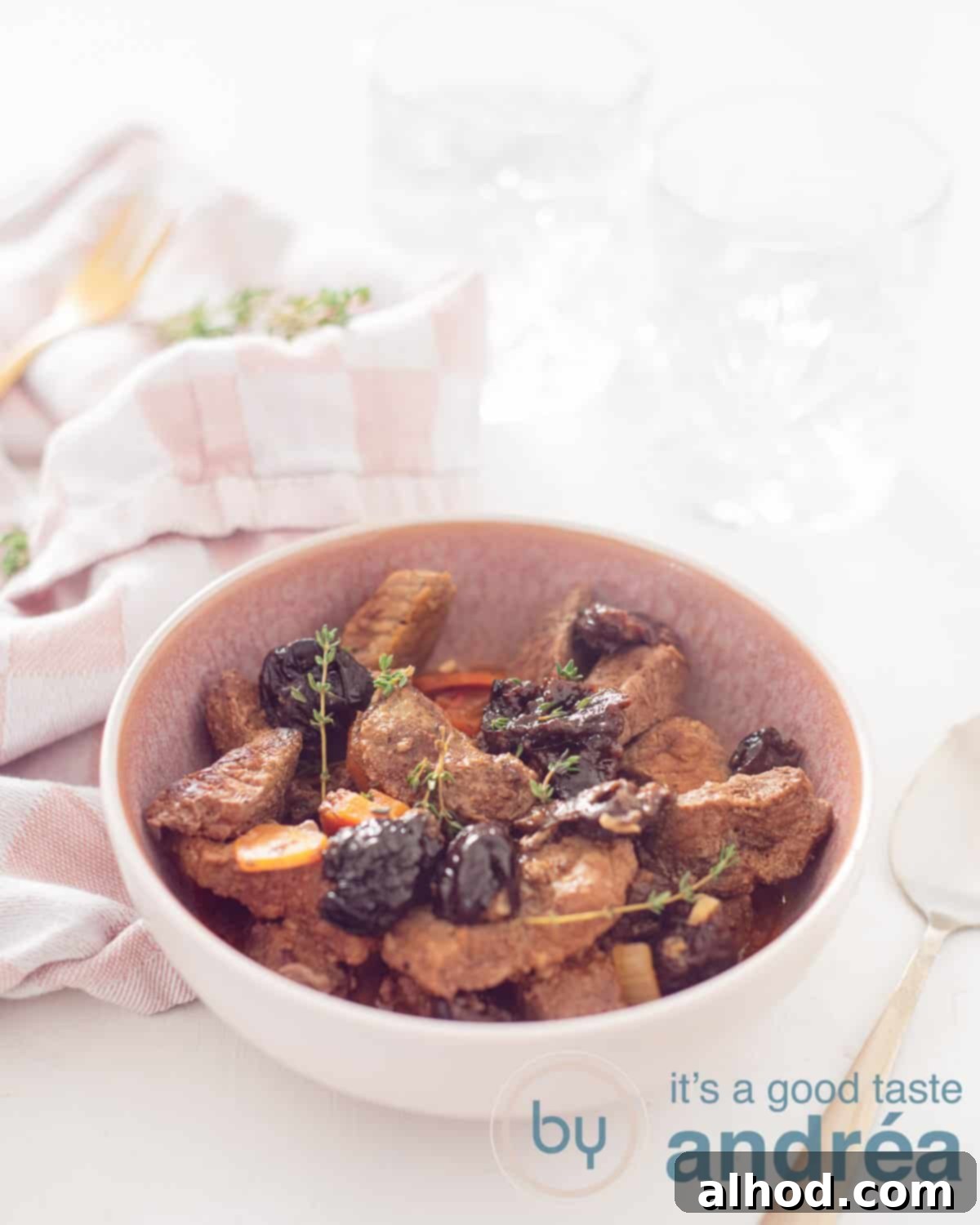 Tender Slow Cooker Plum Pot Roast 2 A height picture with a slow cooker stew with prunes, beef and carrot in the middle. In the back left a pink white tea towel, in the back two glasses on a white background. To the right of the chaal a golden spoon.