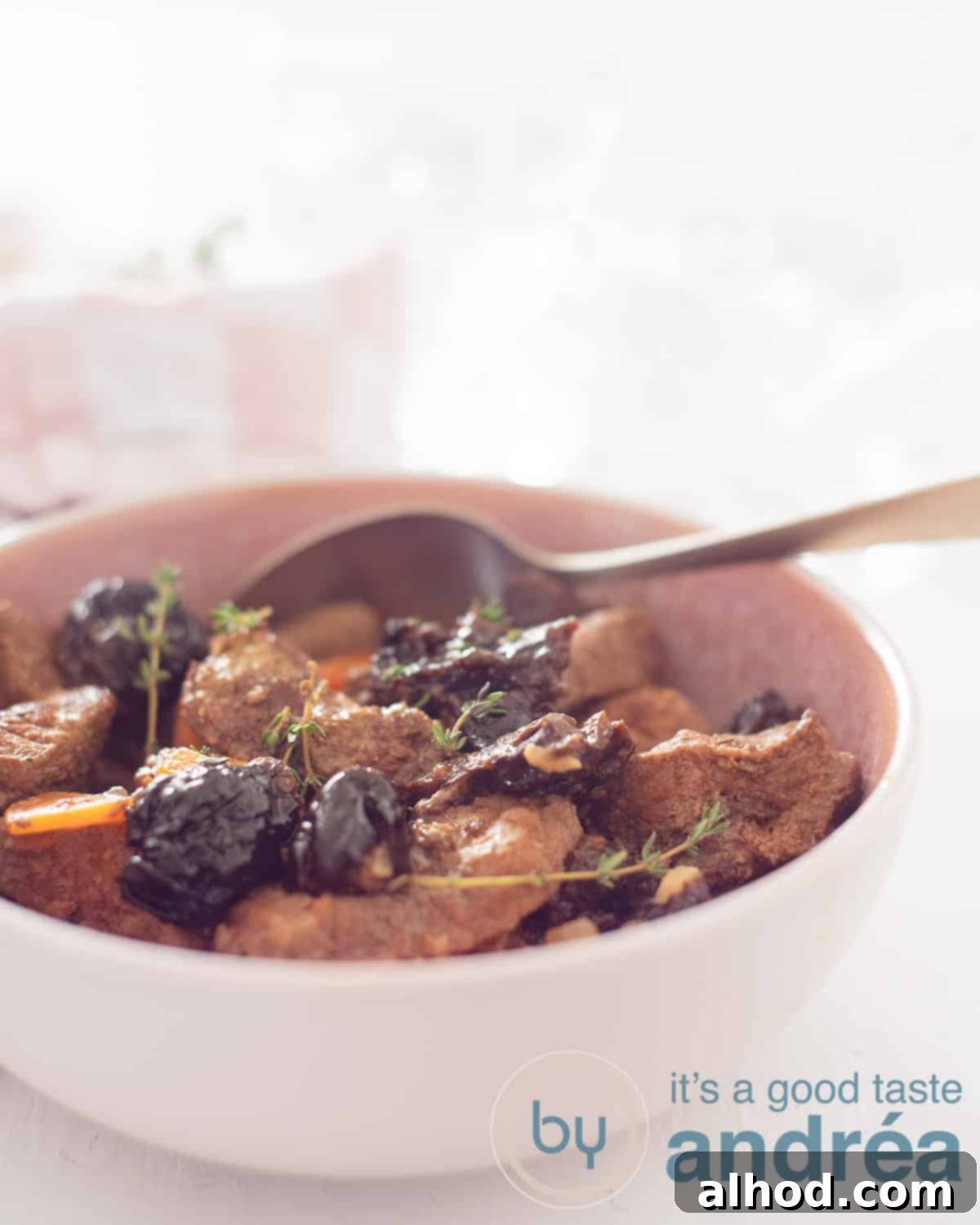 Tender Slow Cooker Plum Pot Roast 5 An high picture of a pink dish filled with beef with prunes and carrot. A golden spoon sticks in it. A white background.