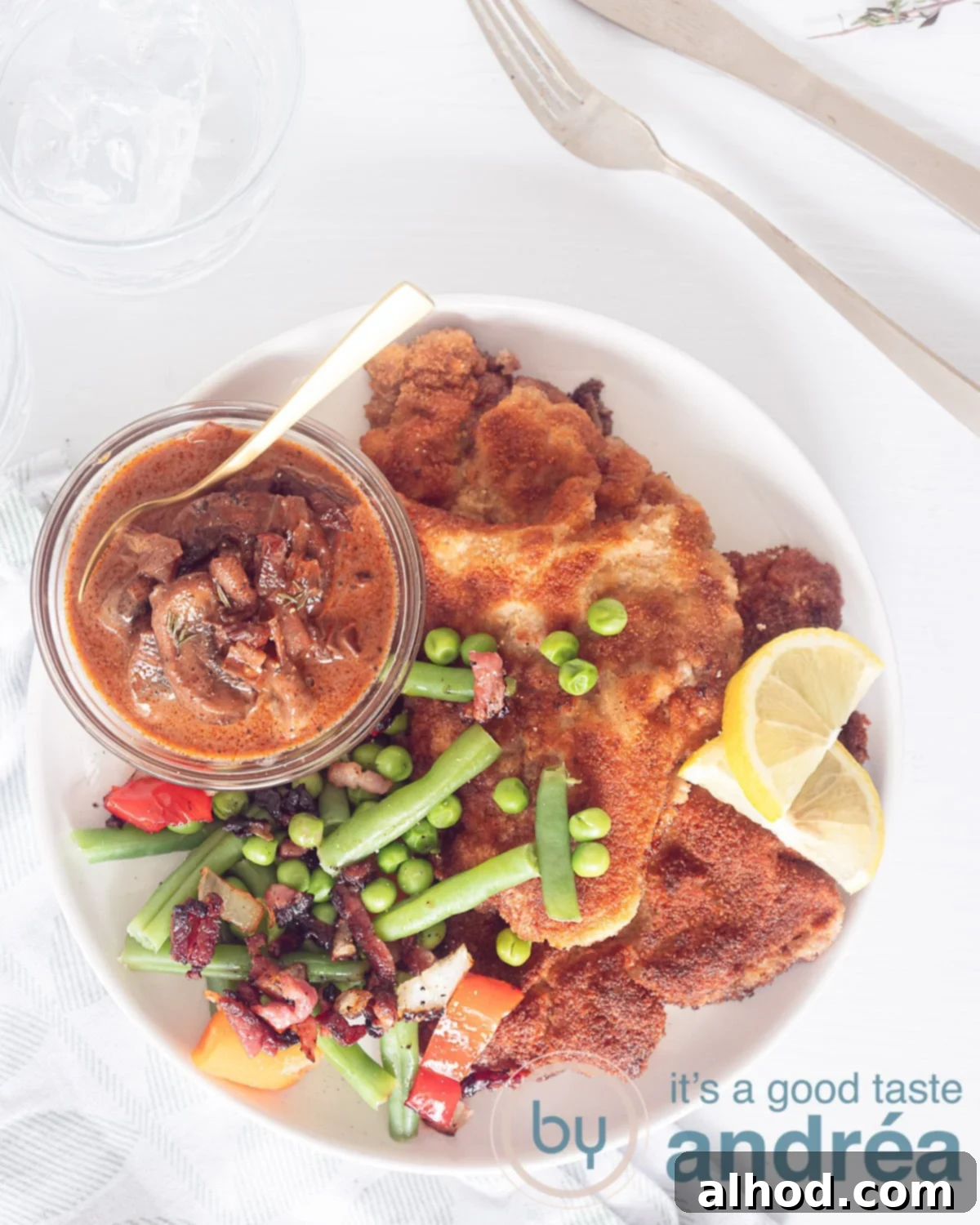 A photo from above of a white plate with schnitzel, fresh vegetables and hunter sauce. Top right gold cutlery. Everything on a white background.