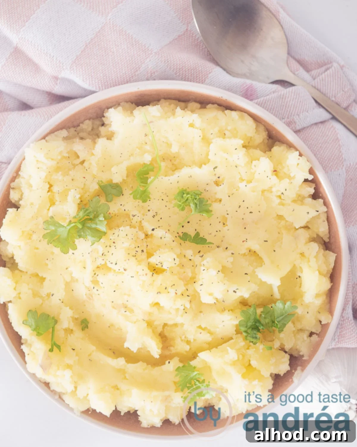 A photo from above with a white background a pink bowl filled with mashed potatoes and garnished with parsley. A white pink tea towel and a golden spoon