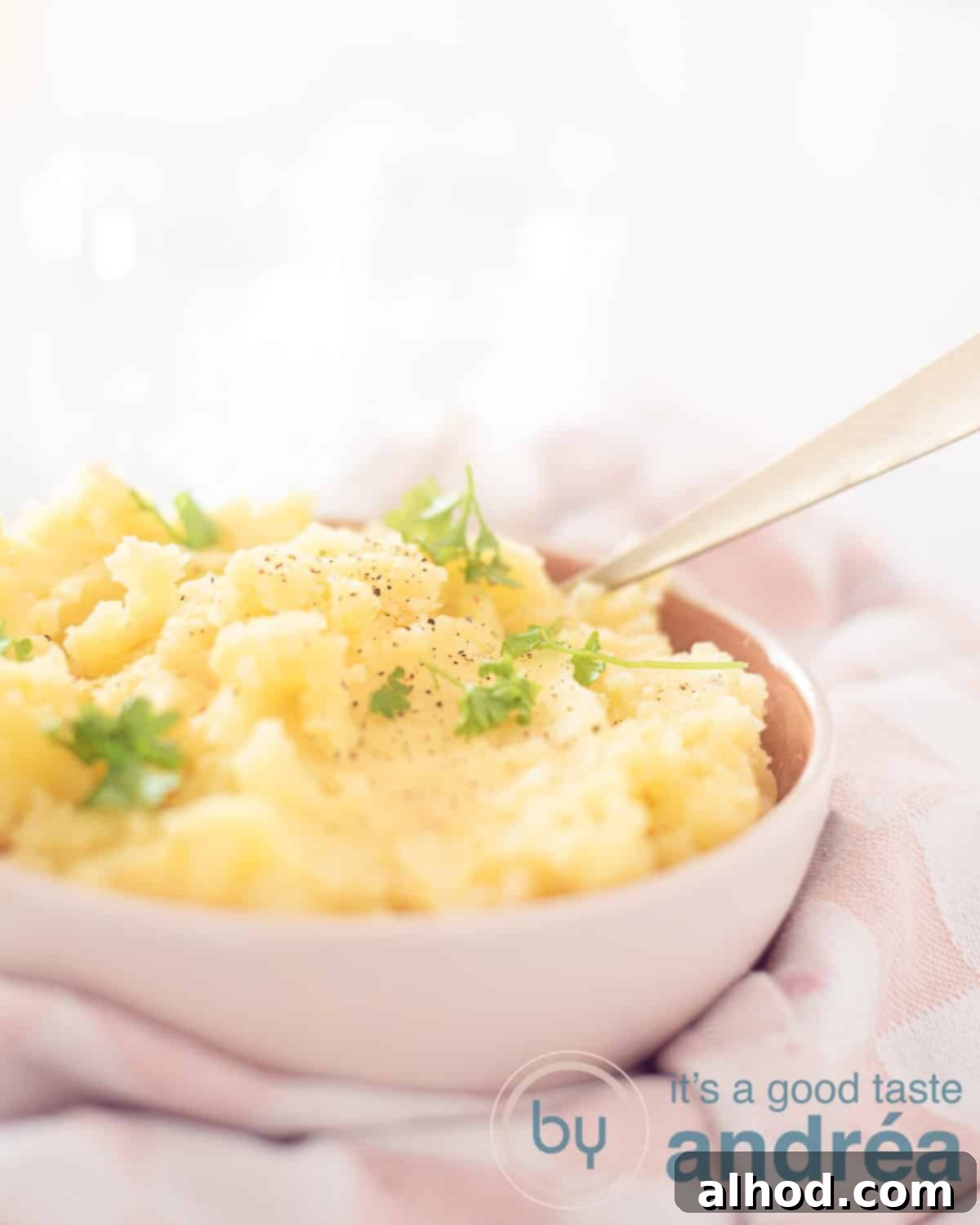 Creamy Garlic & Shallot Mashed Potatoes 5 An aerial view showing part of a pink dish filled with garlic and shallot mashed potatoes, generously garnished with fresh parsley. A golden spoon is stuck into the creamy mash, resting on a pink and white checkered tea towel.