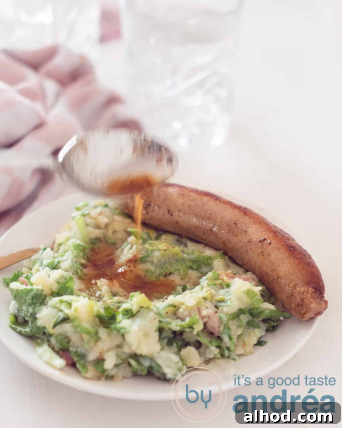Granny's Hearty Endive Sausage Stew 2 A beautifully presented white plate featuring a generous portion of Dutch Endive Stamppot, topped with crispy bacon bits, a juicy pan-fried sausage, and rich gravy poured over. In the soft-focus background, a glass and a pink and white tea towel suggest a cozy, homey setting.