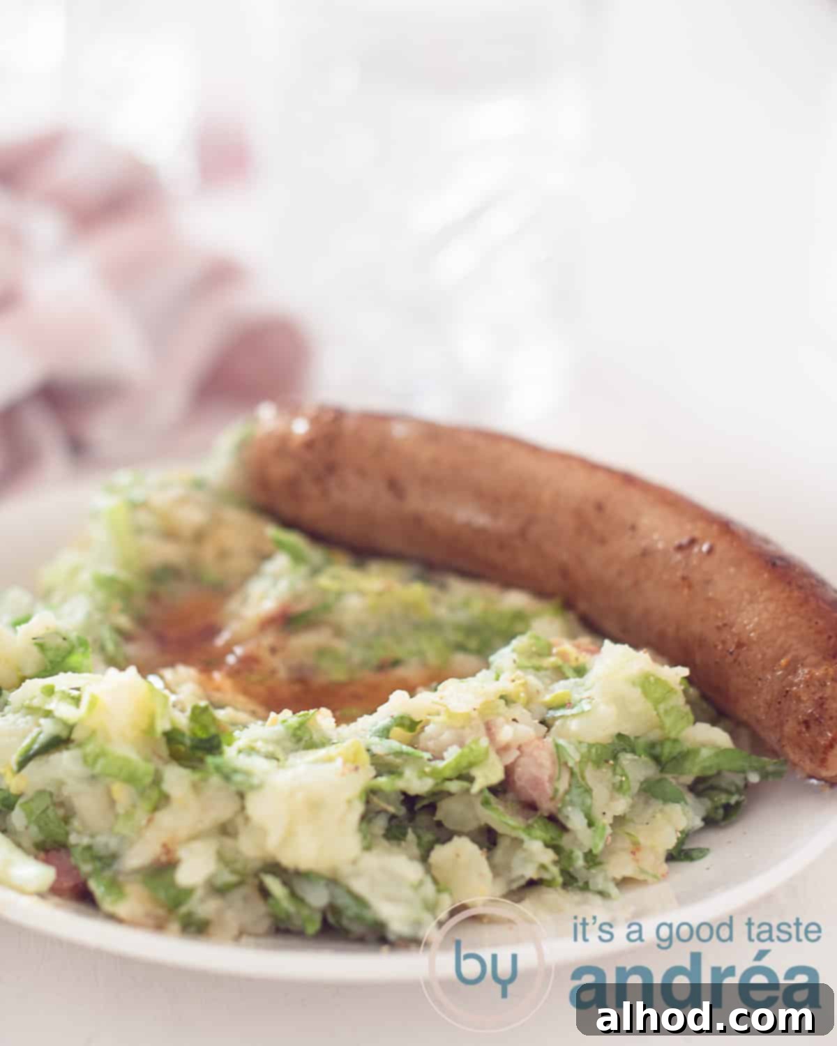 Granny's Hearty Endive Sausage Stew 5 An inviting, elevated shot of a white plate filled with endive stamppot, generously topped with savory gravy and a perfectly cooked sausage. In the background, a elegant glass and a soft pink and white tea towel create a warm, rustic atmosphere.