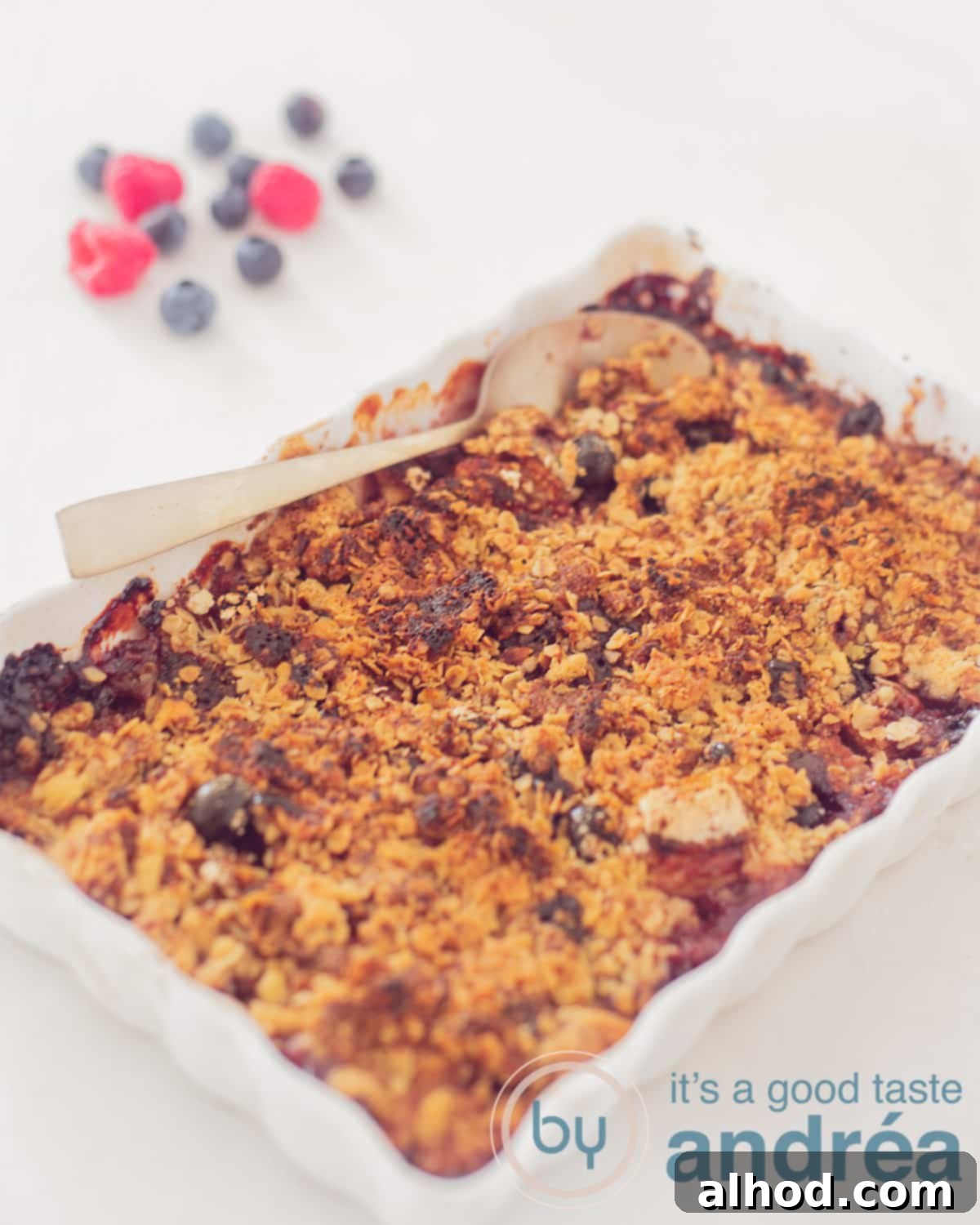 Homestyle Berry Apple Crumble 2 An elevation photo of a white background with an oven dish with apple raspberry crumble. A golden spoon scoops into it. In the background raspberries and berries.
