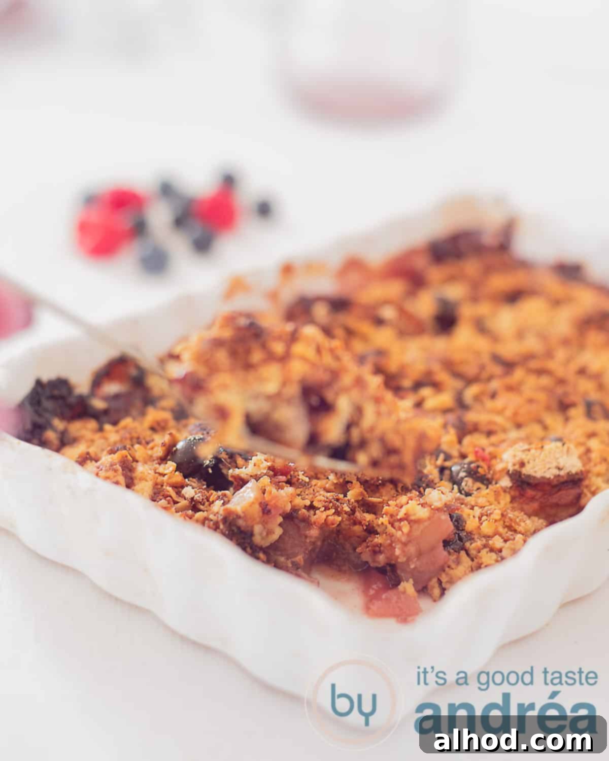 Homestyle Berry Apple Crumble 5 An aerial view showing a white baking dish filled with an apple berry crisp on a white background. Blueberries and raspberries in the background