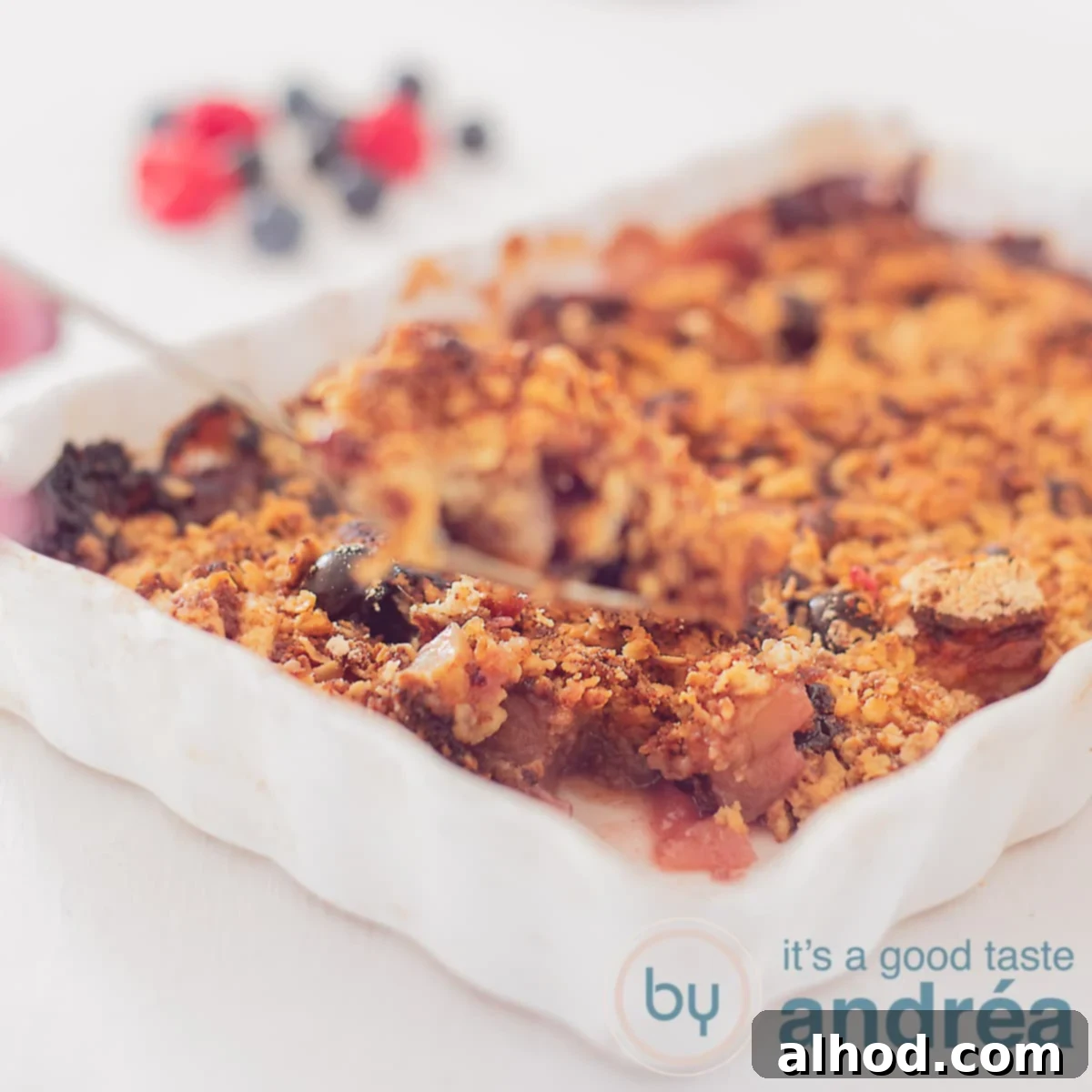Homestyle Berry Apple Crumble 6 A square photo with a white baking dish filled with raspberry oatmeal crumble. A hand holds a spoon with crumble. A white background with berries and raspberries in the background