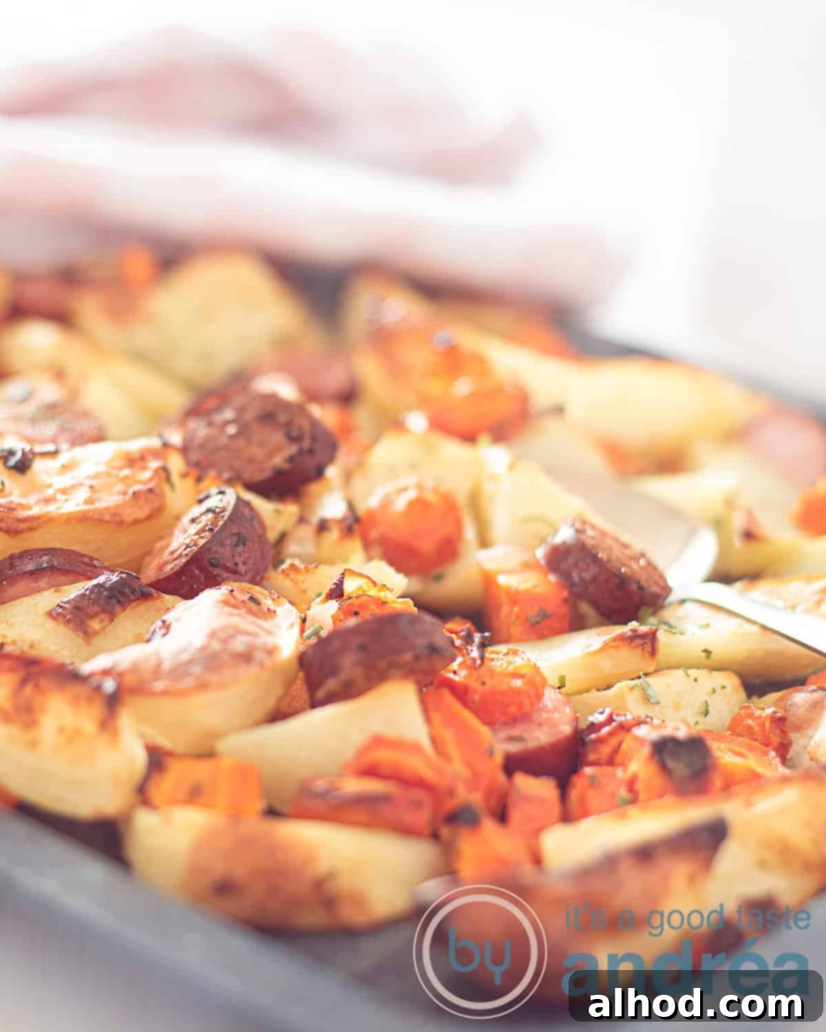 Easy Sheet Pan Sausages 2 An elevation photo with part of a traybake of carrot, smoked sausage and potato. A golden spoon is there. In the background a pink white tea towel.