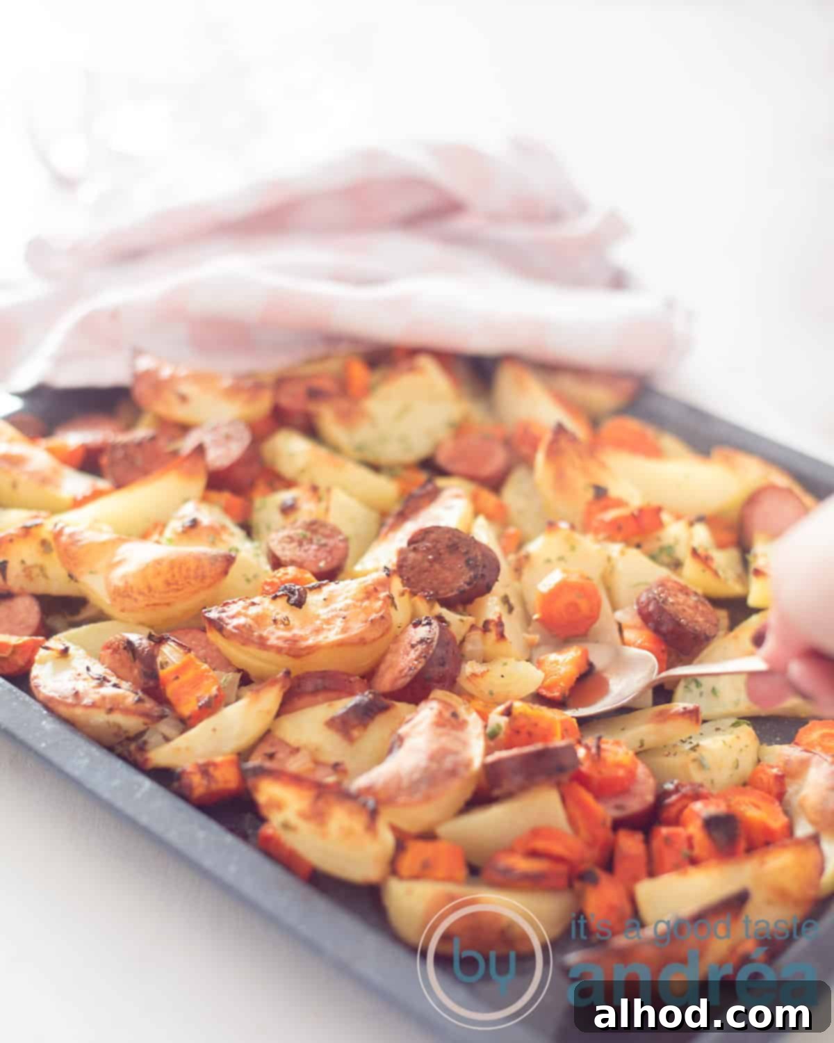 Easy Sheet Pan Sausages 5 An elevation photo with a traybake of potato wedges, carrot and smoked sausage. A spoon scoops out. In the background a pink white tea towel.