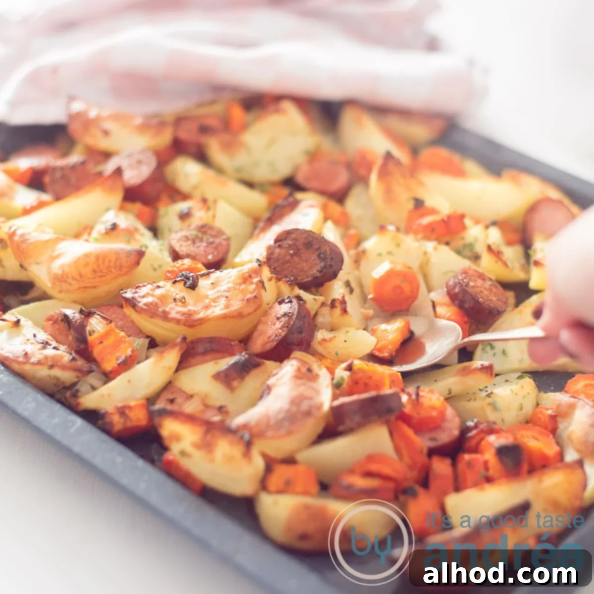 Easy Sheet Pan Sausages 6 A square shot of a griddle filled with roast potato, carrot and smoked sausage. A spoon takes out a scoop. In the background a pink white tea towel.