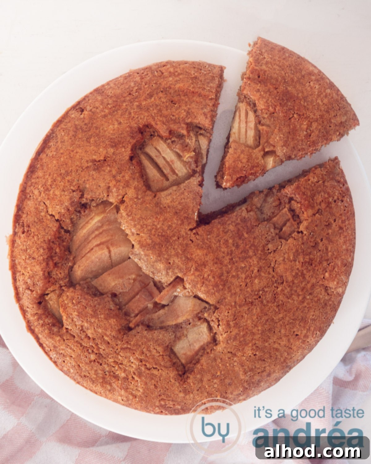 A beautifully presented slice of moist pear cake with olive oil, showcasing the juicy pear pieces within, set on a white cake tray with a cut-out section slightly loose. The background is white.