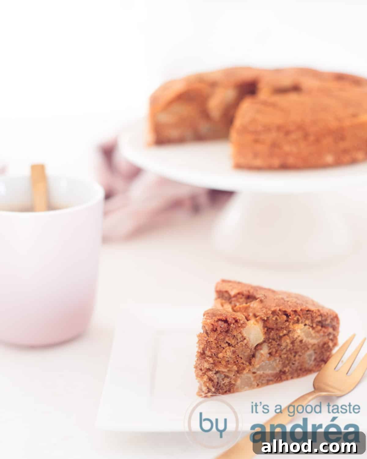 An inviting overhead shot of a slice of warm, moist pear cake with olive oil in the foreground, with the remaining cake on a white cake stand in the background, alongside a pink coffee cup.