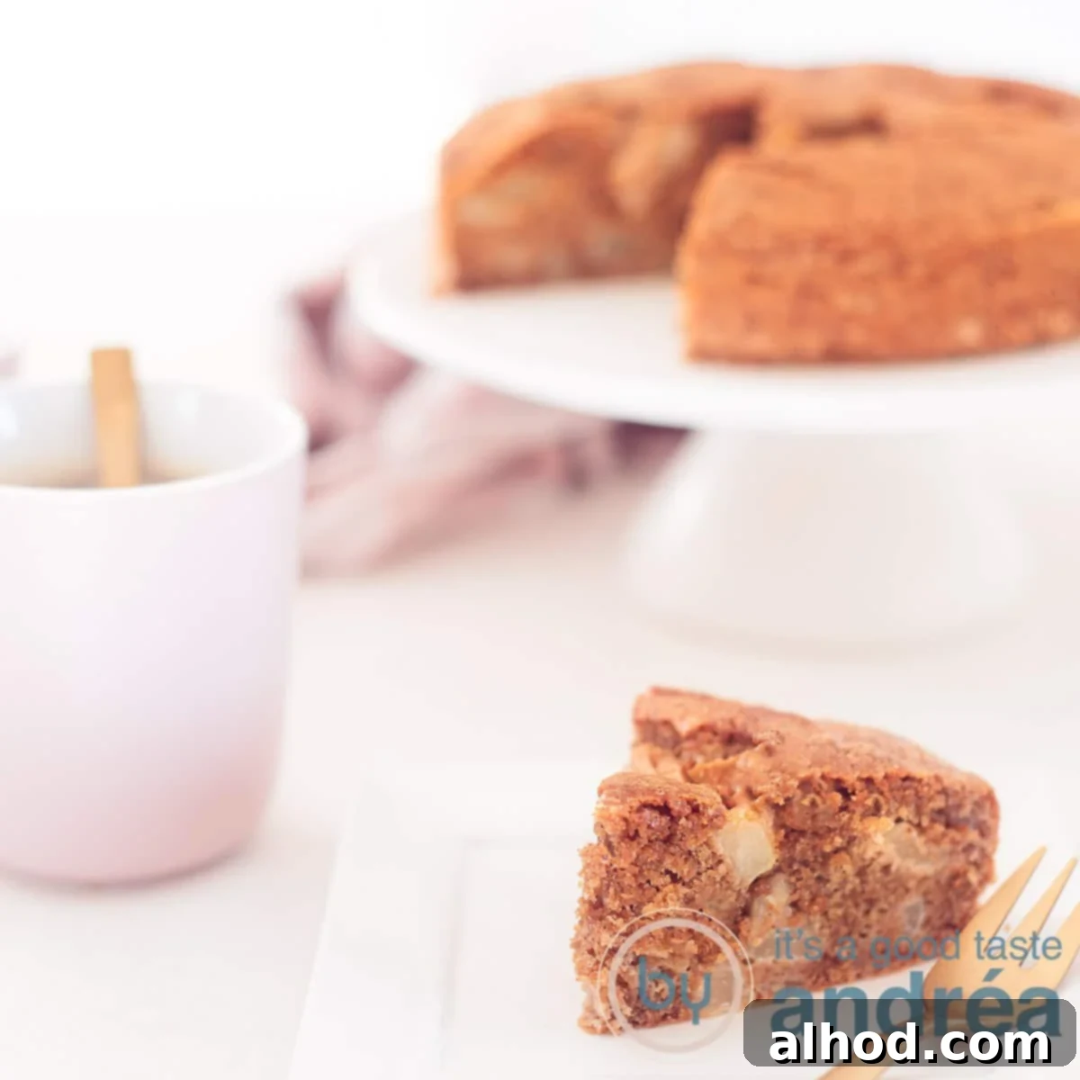 A square photo with a slice of moist pear cake in the foreground and a cake stand with the rest of the cake in the background. A pink cup of coffee is visible on the left.