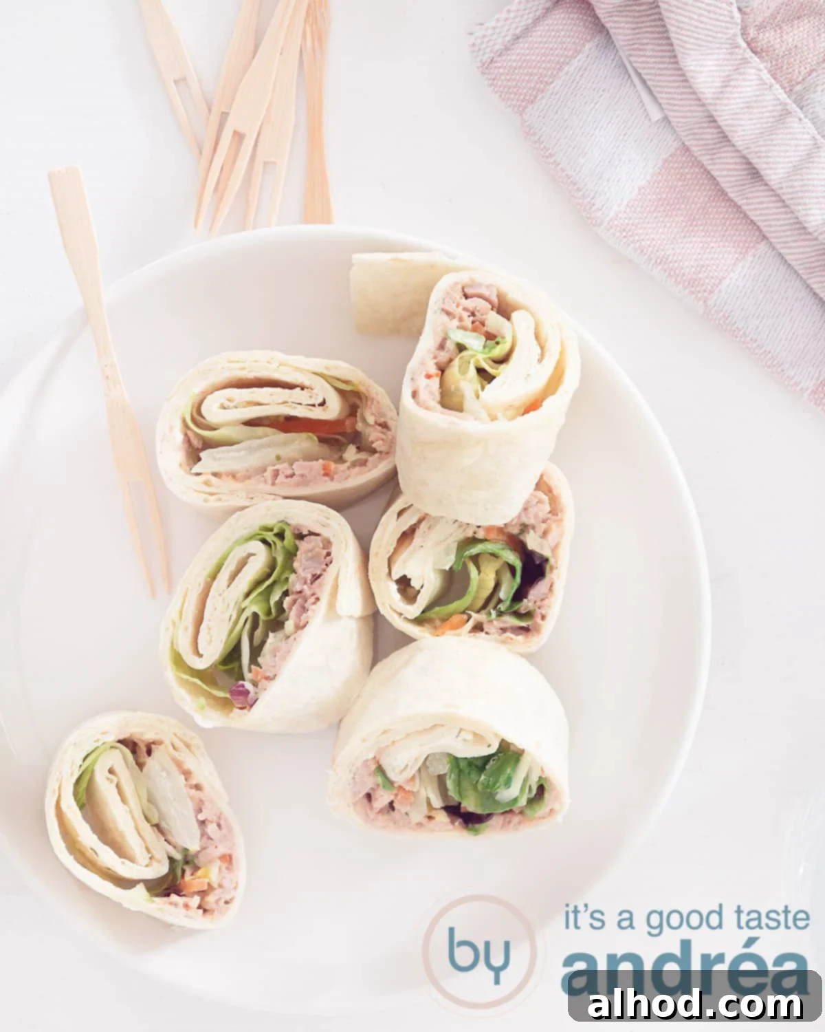 Creamy Tuna Spirals 2 A photo from above. Bottom left a white plate with six wraps with tuna and salad. Sticks are there. A white background. Top right a pink white tea towel.