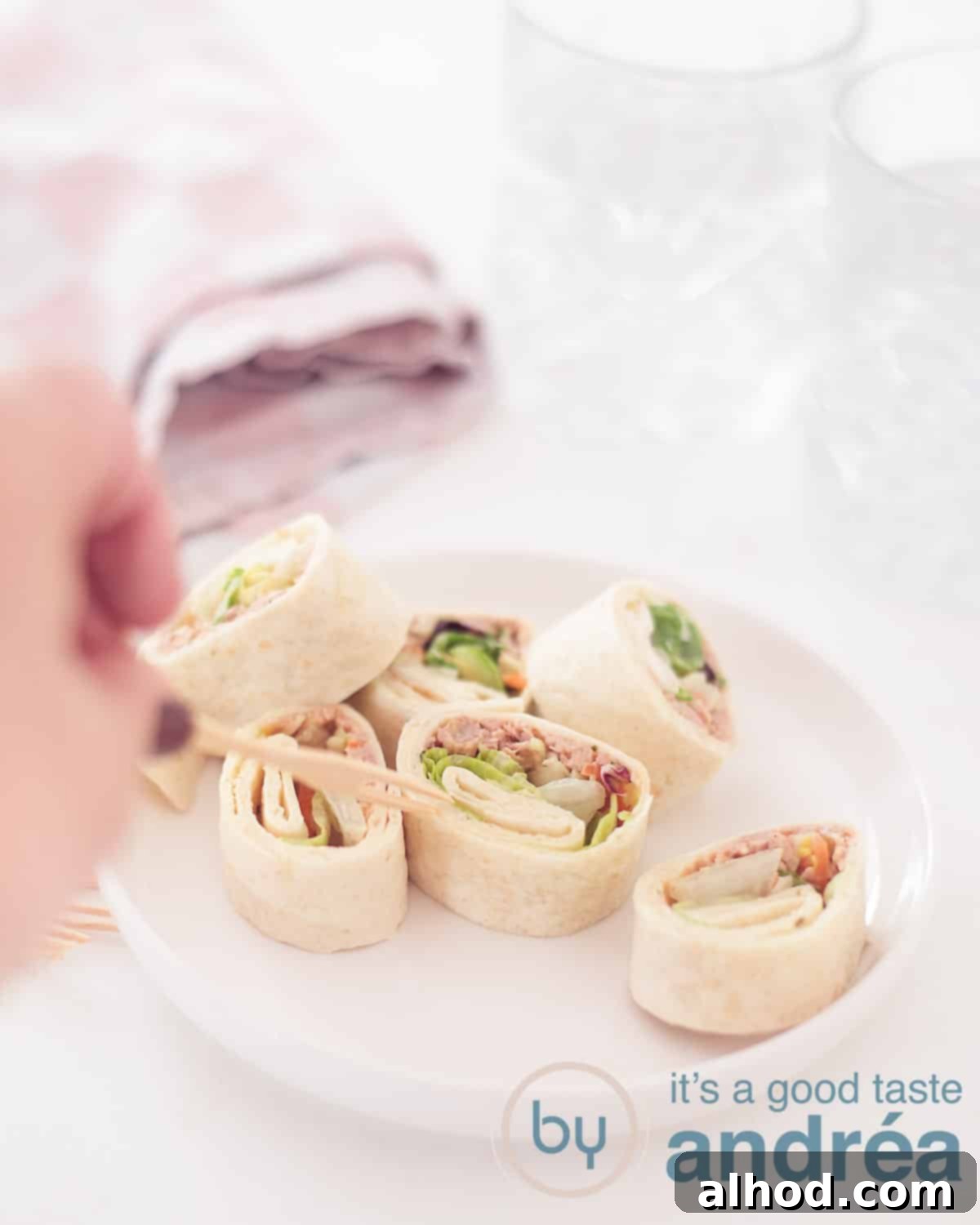 Creamy Tuna Spirals 5 An elevation photo with a white plate with various tuna salad wrap snacks. A hand pricks a bite. A white background with two glasses and a pink white tea towel in the background.
