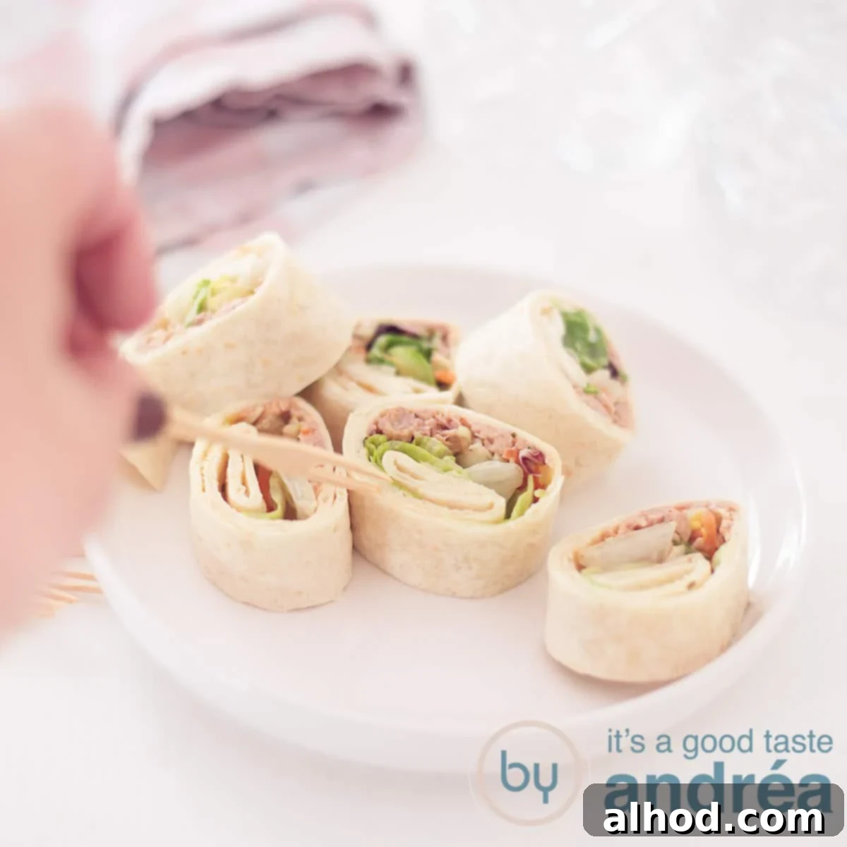 Creamy Tuna Spirals 6 A square photo with a plate full of wrap snacks with tuna. A hand pricks a wrap snack. A white background.
