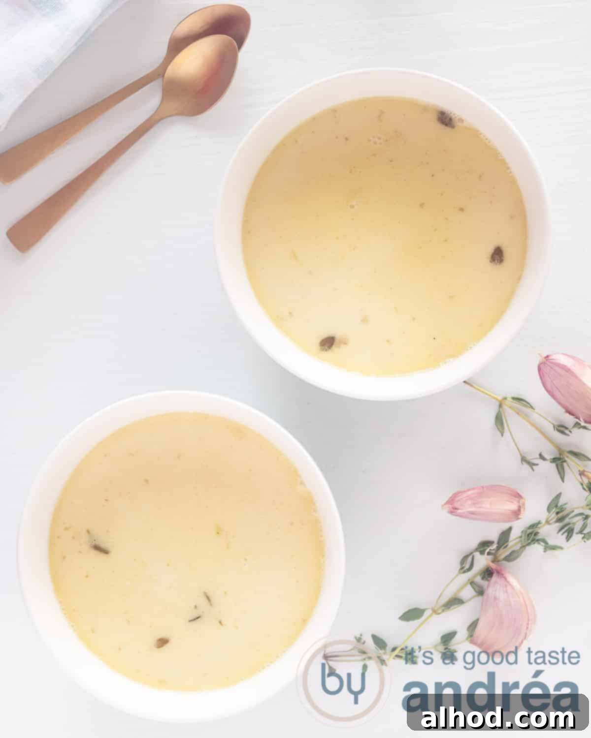 Velvety Roasted Garlic Soup 2 Two bowls filled with creamy roasted garlic soup on a white surface, garnished with fresh thyme. Golden spoons and garlic cloves are scattered nearby.