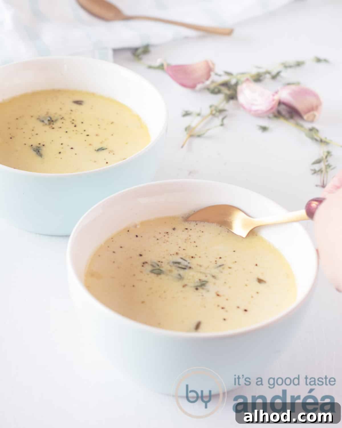 Velvety Roasted Garlic Soup 5 An overhead shot of two blue bowls of roasted garlic soup. A golden spoon is dipping into one bowl. Fresh thyme sprigs and garlic cloves are in the soft-focus white background.