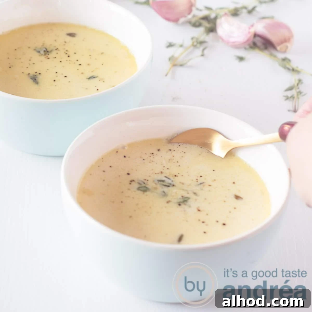 Velvety Roasted Garlic Soup 6 A square photo with two blue bowls filled with soup made from garlic, wine and broth. A hand takes a bite with a golden spoon. Garlic and thyme (fresh) in the background).