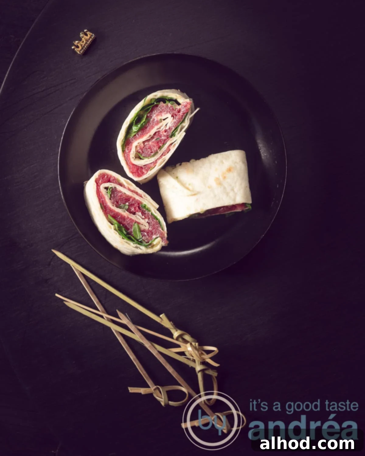 Classic Beef Carpaccio Pinwheels 2 A height photo in the top center of a black plate with three carpaccio wraps. Tapas sticks at the bottom.