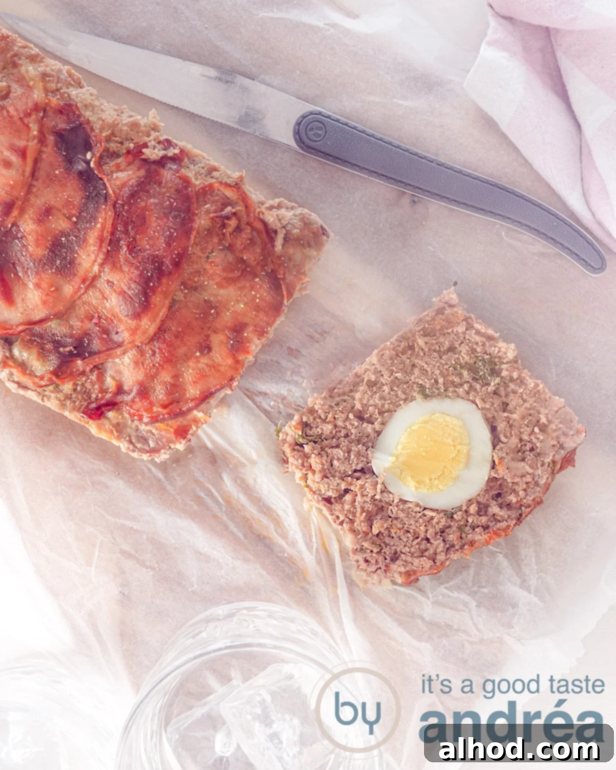 German Mock Hare Meatloaf 2 A photo from above of minced meat in the form of a loaf filled with egg and a topping of crispy bacon. At the top a knife and a pink white tea towel. Located on a baking paper.