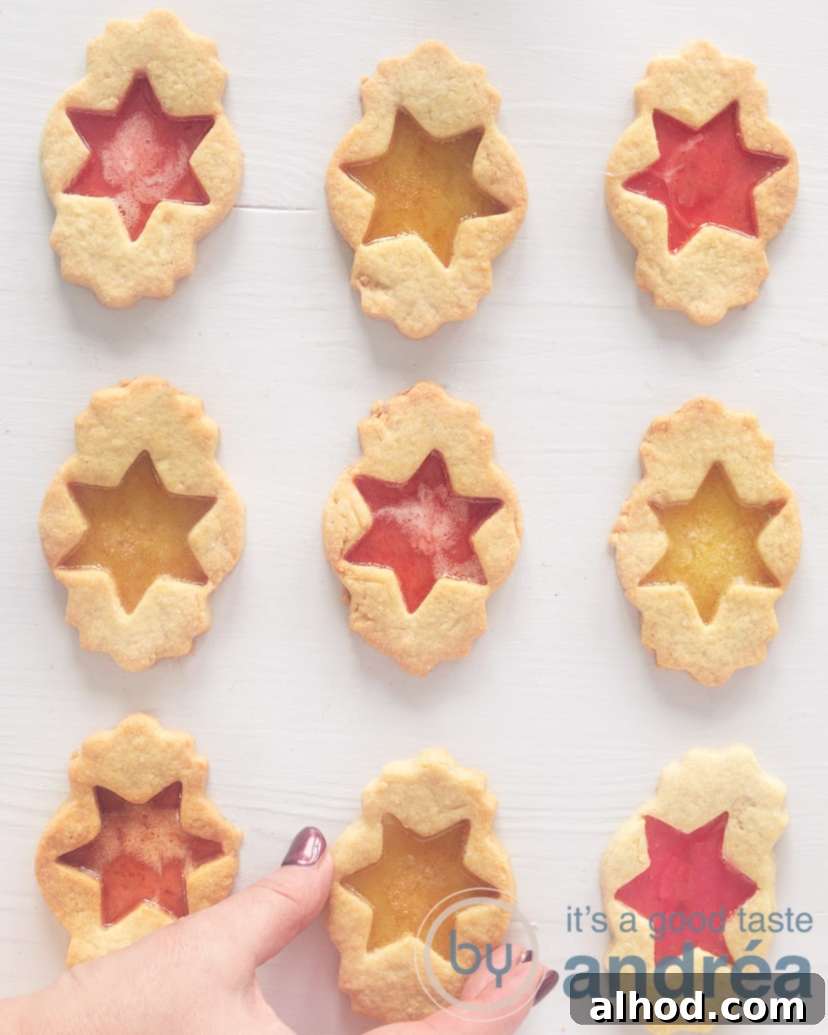 Jeweled Window Cookies 2 A stunning overhead shot of nine stained glass biscuits, featuring alternating red and yellow candy fillings arranged in a square. A hand reaches for the cookie at the bottom center. The cookies are set against a pristine white background, highlighting their festive colors.