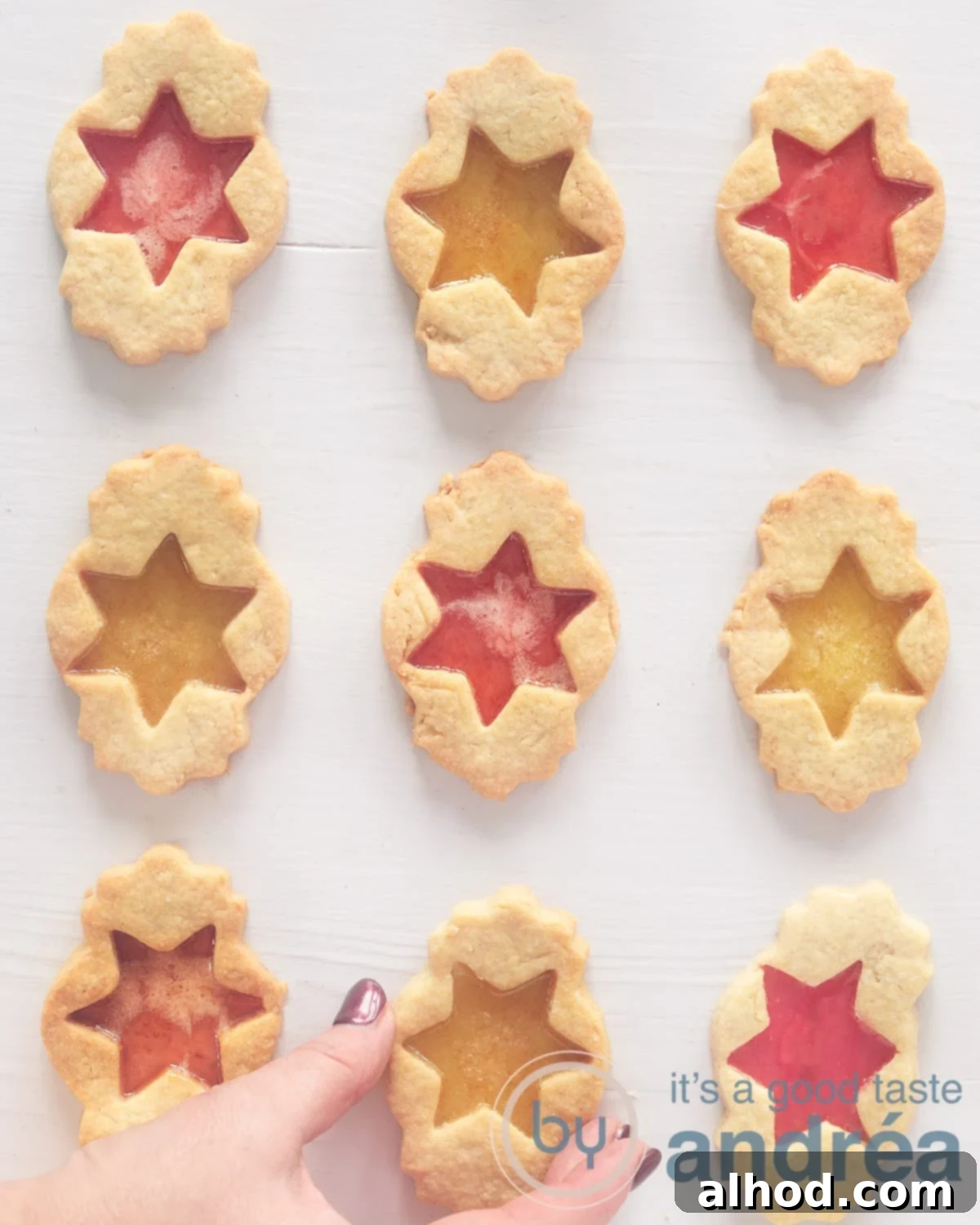 A photo from above of 9 stained glass biscuits with a red and yellow filling alternated in a square. The biscuit in the middle of the bottom is grasped. A white background.