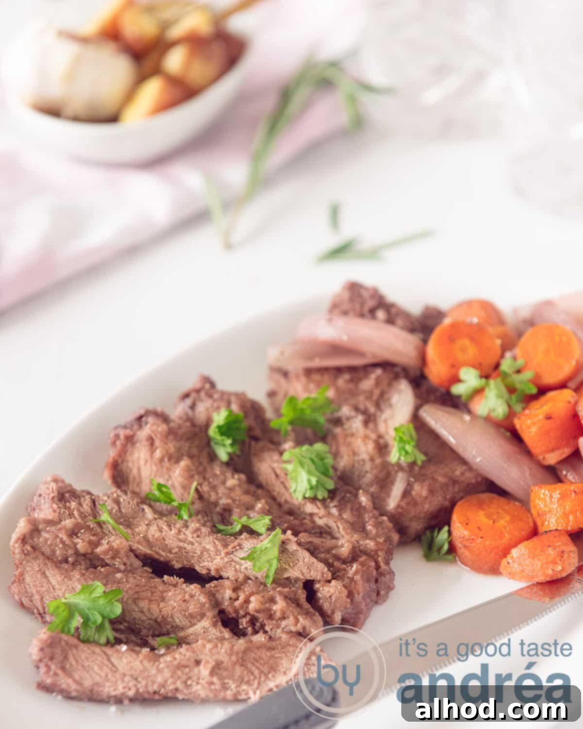 Succulent Roasted Sirloin with Deep Red Wine Sauce 5 A dish showcasing the stewed sirloin steak, tender shallots, and carrots in a rich red wine sauce. In the background, a bowl of crispy roasted potatoes with roasted garlic on a white backdrop, ready to be served.
