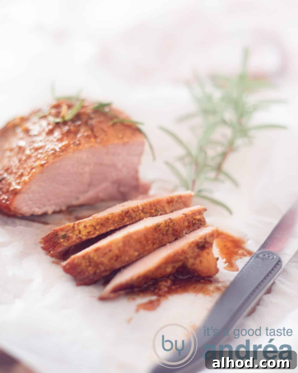 A perfectly cooked ham with a glistening maple glaze, three slices already carved, resting on white parchment paper. A sprig of fresh rosemary and a steak knife are positioned nearby, enhancing the visual appeal.