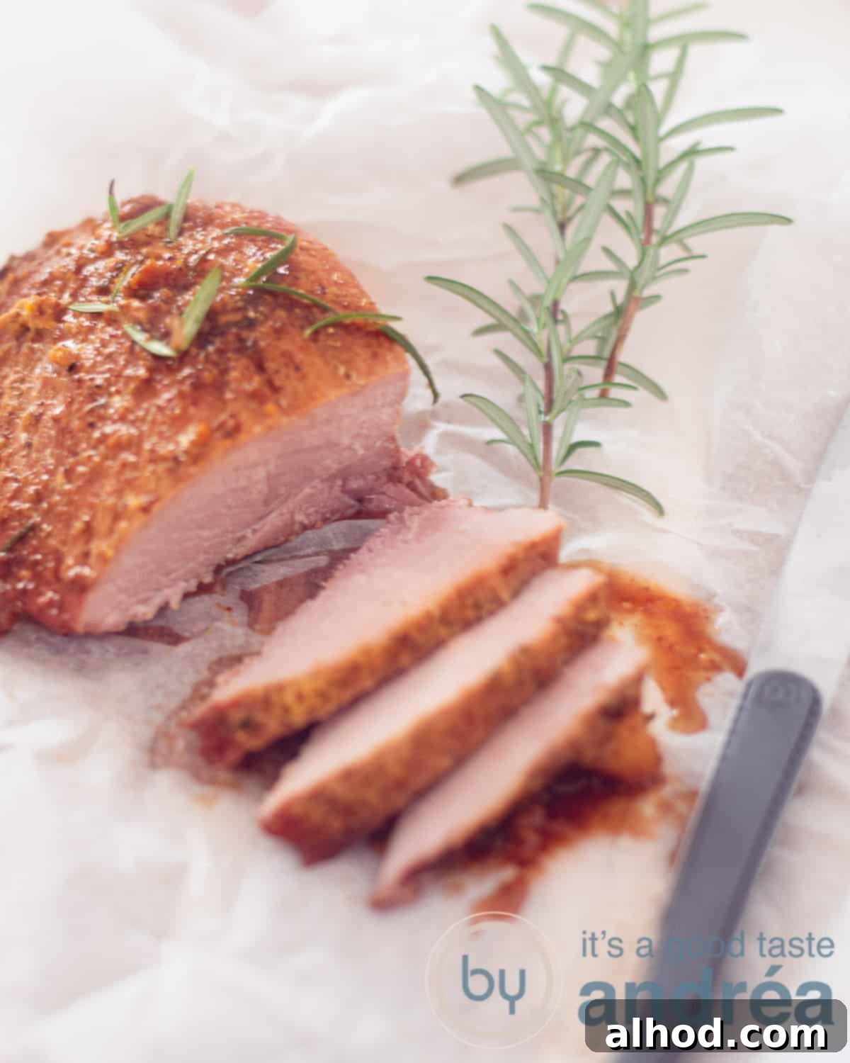 An appetizing elevation photo of a maple glazed ham, three slices expertly cut, showcasing the tender interior and golden-brown glaze. It rests on white parchment paper, flanked by a steak knife on the right and a sprig of rosemary at the top.