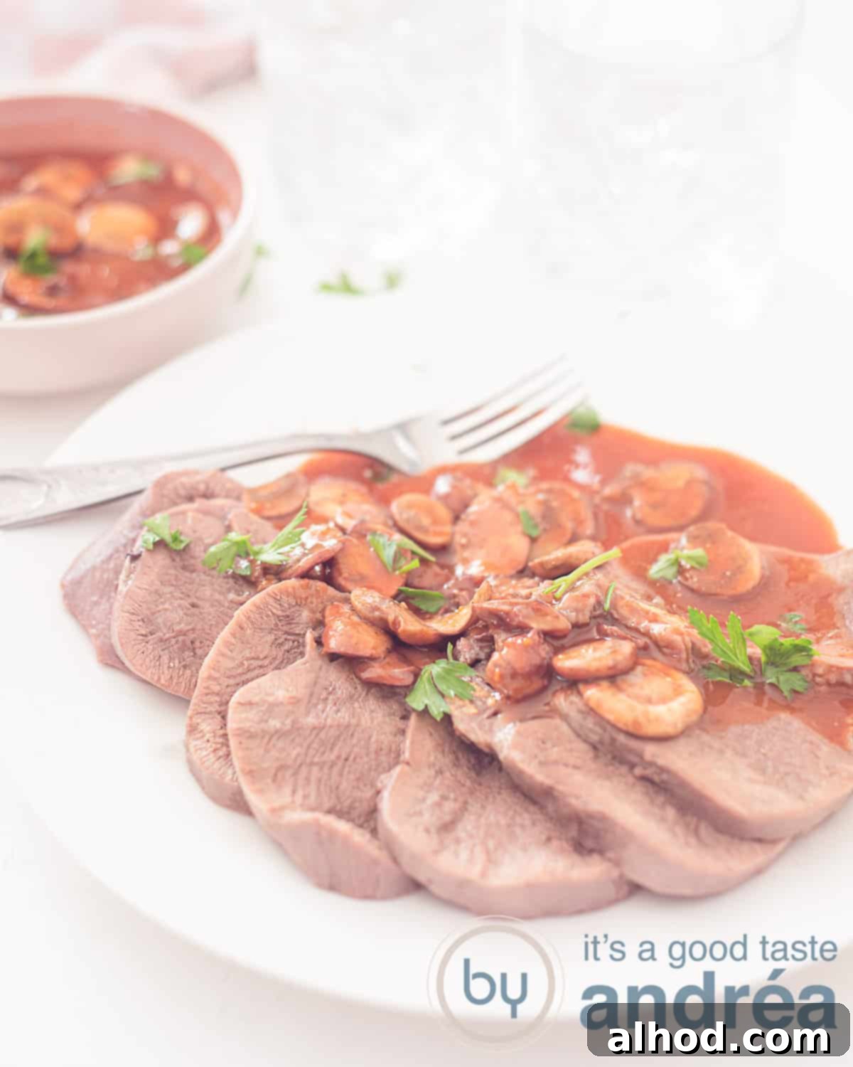 Tender slices of braised beef tongue, arranged elegantly on a white plate and generously topped with a rich, glossy Madeira sauce, garnished with fresh parsley. A bowl of additional sauce rests in the background, all on a pristine white surface.