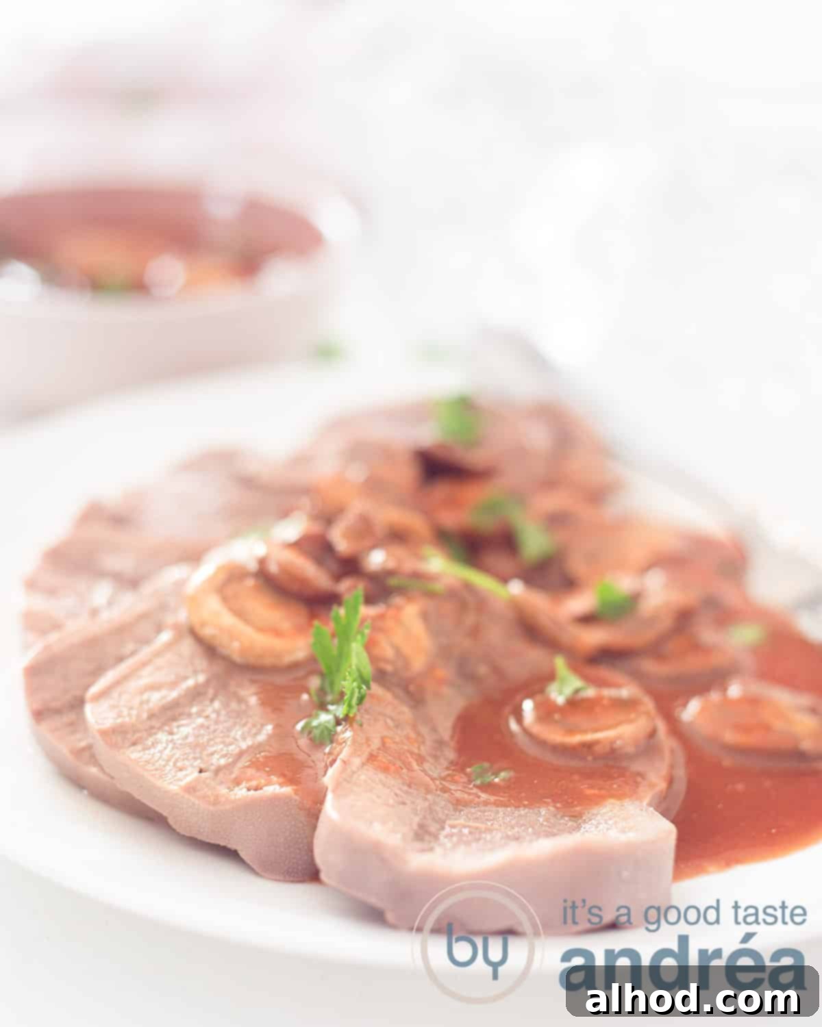 An appetizing close-up of a white dish featuring tender slices of beef tongue generously covered with glossy Madeira sauce, beautifully garnished. A bowl of extra sauce is blurred in the background, all set against a clean white backdrop.