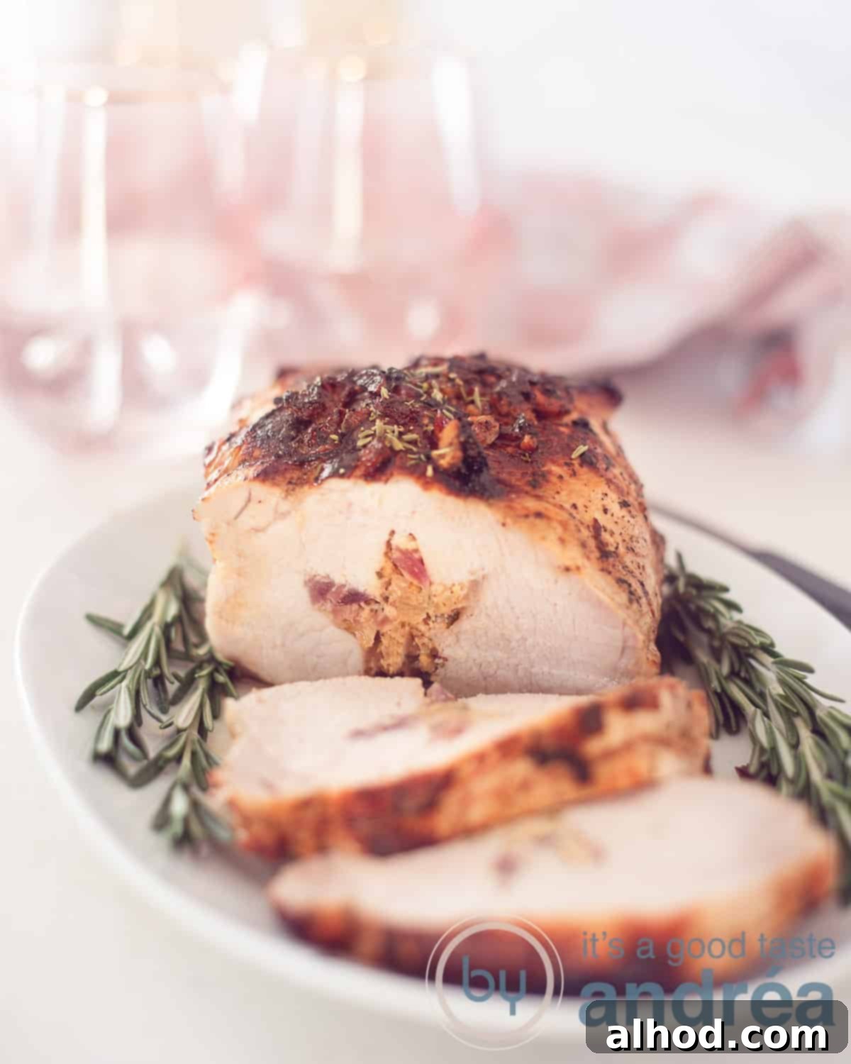 Traditional Dutch Rolled Roast 5 An elevation photo with a small plate of rosemary, roulade with a filling of onion, hazelnut, mustard and honey. A tea towel with pink white and two glasses in the background.