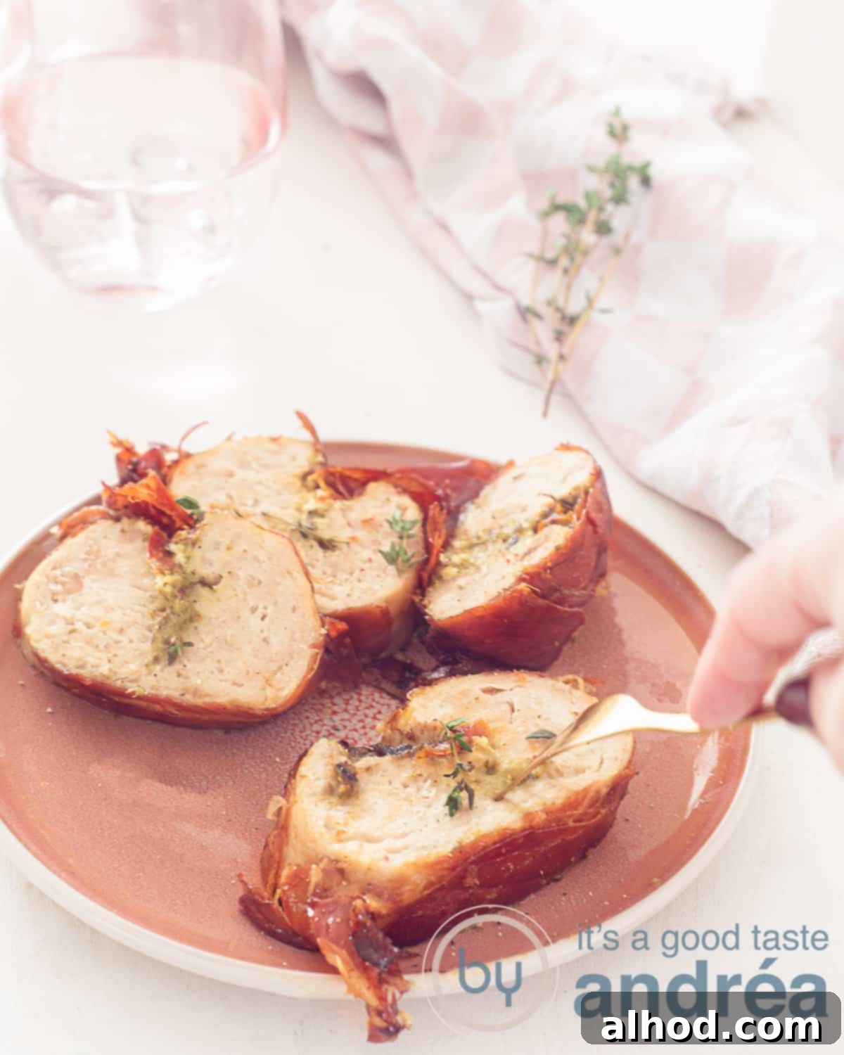 Dutch Chicken Roulade 2 An elevation photo with a pink plate filled with chicken roulades and wrapped in prosciutto. A fork pierces it. A white background.
